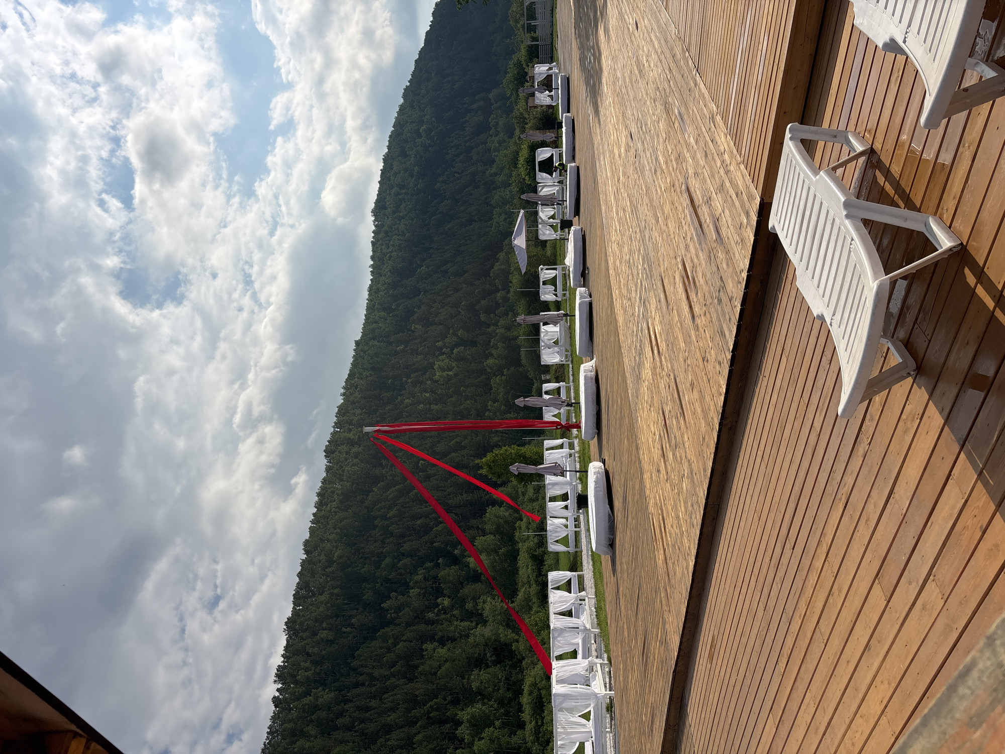 The image depicts an outdoor recreational area at a resort or beach club, likely in or near Дрокино, Russia, given the surrounding lush, forested hills. The foreground is dominated by a large, light brown wooden deck, appearing slightly damp. Two white plastic lounge chairs are positioned on the deck, one fully visible and the other partially.
In the mid-ground, a line of white cabanas with draped fabric sides are set up on a grassy area. These cabanas appear to be for relaxation or seating. Several large, circular, white-covered daybeds are also visible in this area, some with umbrellas. A tall pole stands prominently in the center, from which vibrant red fabric streamers extend outwards, creating a decorative element. More umbrellas and cabanas can be seen further back, suggesting an extensive leisure facility.
The background features a dense, dark green forest covering a steeply sloped hill. The sky is partly cloudy, with a mix of white and grey clouds against a pale blue sky, indicating fair weather. The overall impression is one of a well-maintained outdoor entertainment space designed for guests to relax and enjoy the natural surroundings. There are no people visible in the image.