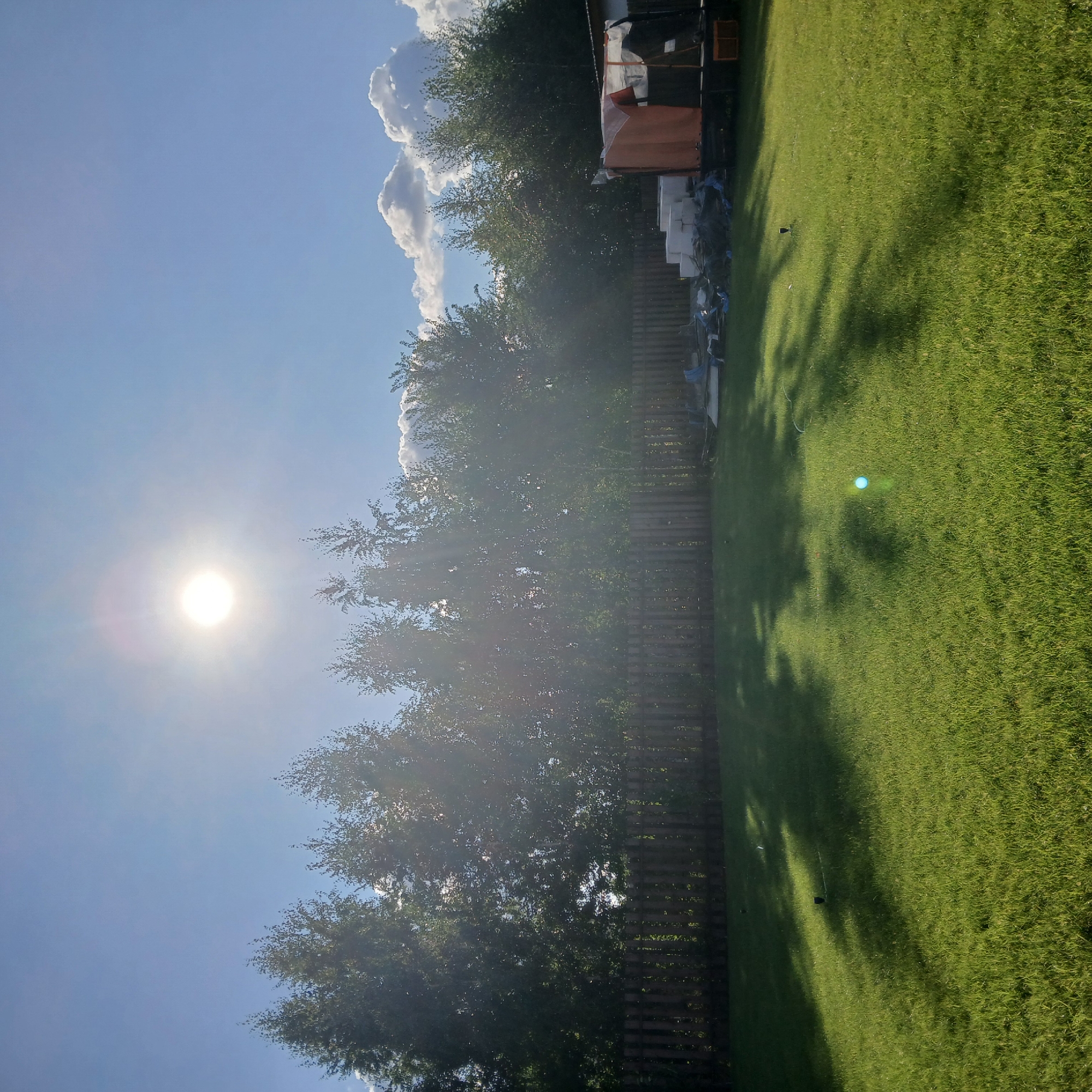 The image depicts a backyard scene on a sunny day with the sun high in the sky. A line of trees forms a dense backdrop behind a wooden fence. In the foreground, a well-maintained green lawn is visible, with sprinklers casting shadows across it. On the right side of the frame, near the fence, are several items including white building blocks, some tarps, and a partially covered structure. The sky is a clear blue with a few white, puffy clouds on the right horizon. The overall impression is a suburban or rural backyard during daylight hours. The location is not specified within the city of Samarsky, Russia.
