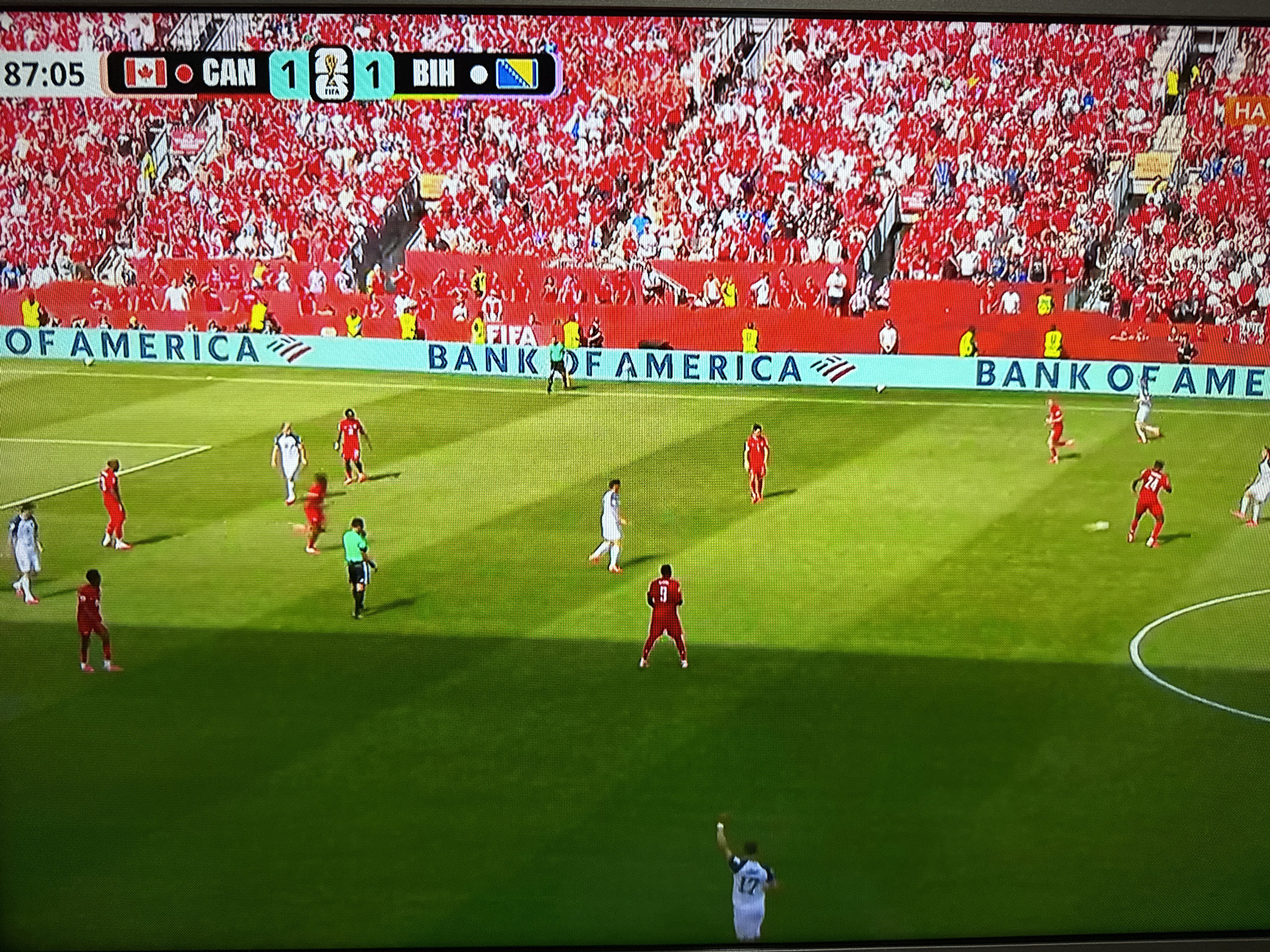 The image shows a soccer match being played in a stadium with a large crowd in the stands, all wearing red. A scoreboard at the top of the screen indicates the time is 87:05 and the score is CAN 1, BIH 1. The game is sponsored by Bank of America. Players in red and white uniforms are visible on the green field, with some players in red jerseys and white shorts, and others in white jerseys and black shorts. One player in a red jersey with the number 9 is standing in the middle of the field. The scene is illuminated by stadium lights, casting shadows on the grass. The photograph is taken from a slightly elevated angle, looking down at the field and the crowd. There is no indication of the city of Targovishte, Bulgaria being relevant to this image.