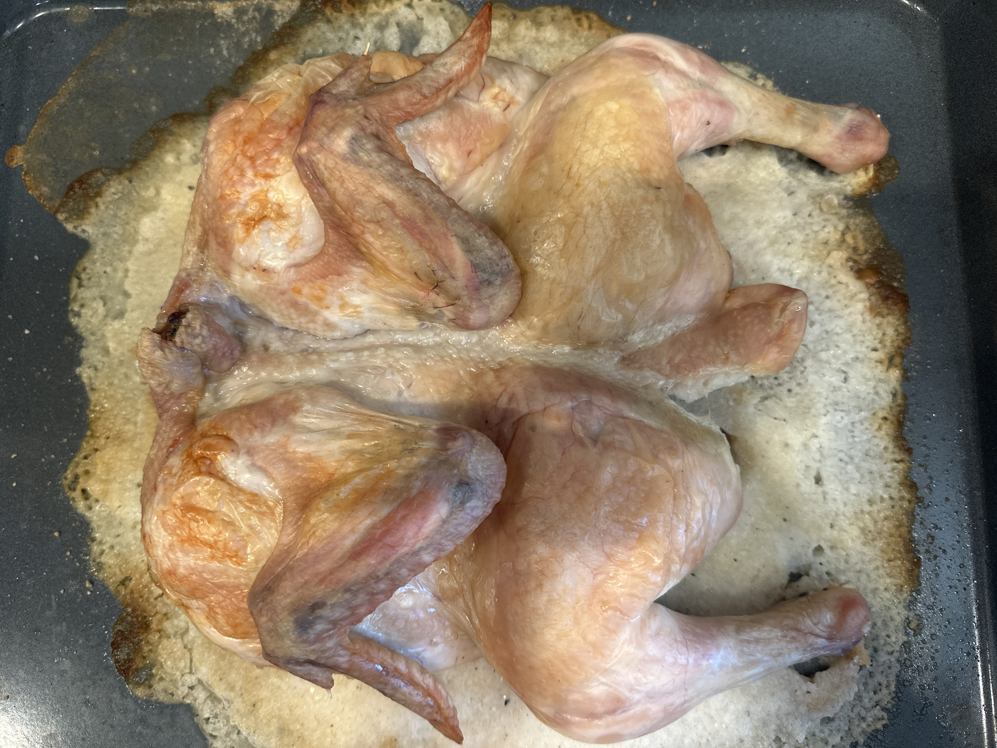 The image shows a whole, raw chicken that has been butterflied and is resting on a baking tray. The chicken's skin is pale pink and slightly translucent, with some areas of orange-brown discoloration, particularly around the wings and breast. The legs are spread apart, and the wings are tucked underneath the body. The baking tray appears to be dark gray and has a white, crumbly substance around the chicken, which may be salt or a seasoning. There are also some areas of browning and charring on the tray, suggesting it has been used for cooking. The lighting is overhead, casting soft shadows on the chicken. There is no indication of the location within the city of Minsk, Belarus.