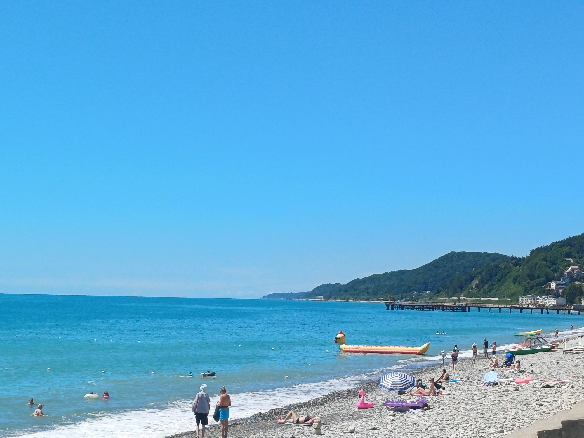 This is an eye-level, medium shot of a pebble beach on a sunny day. The sky is clear and bright blue. The sea is a vibrant turquoise color, with gentle waves lapping at the shore. In the water, several people are swimming and wading. Some are using inflatable rings. A long, yellow inflatable boat shaped like a banana is floating in the sea.
On the beach, people are sunbathing, sitting under umbrellas, and walking. There are beach chairs and umbrellas set up. A pink inflatable flamingo and a purple inflatable raft are visible on the pebbles. Two people, a man and a woman, are walking towards the water. A woman is lying on a towel on the beach.
In the background, a long pier extends out into the sea. Beyond the pier, lush green hills rise up, with buildings scattered on their slopes. The scene suggests a popular tourist destination, likely the city of Sochi, Russia, known for its Black Sea coast and mountainous terrain.