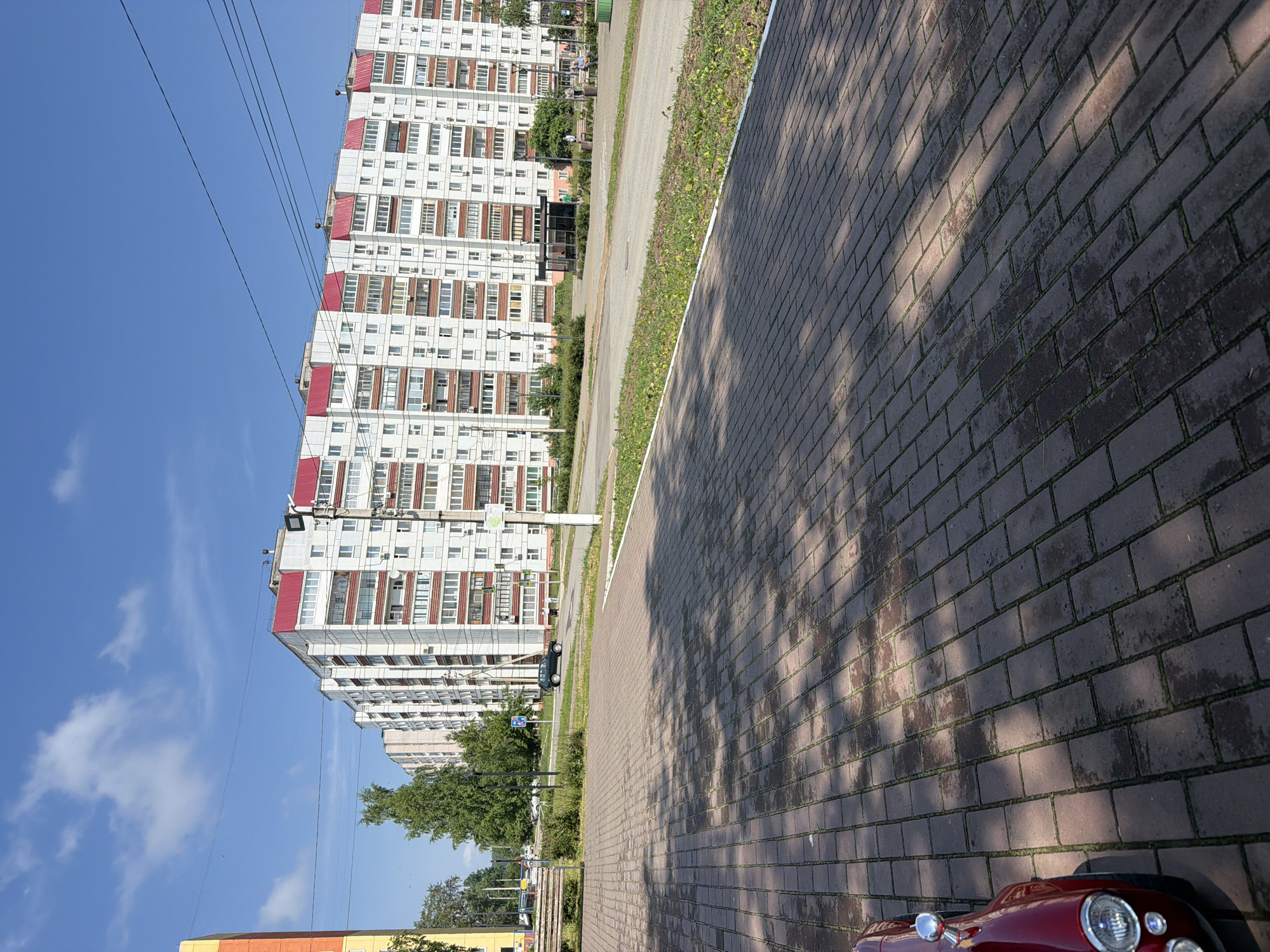 In Chaikovsky, Russia, on a bright, sunny day with a clear blue sky and scattered clouds, a row of multi-story apartment buildings with red roofs dominates the background. The buildings are white with red balconies and windows. In the foreground, a red car is partially visible on a paved walkway. The walkway is made of red brick pavers, and shadows from trees are cast across it. A bench and some greenery are also present in the mid-ground, along with a bus stop shelter and several people. A street runs parallel to the buildings, with a few cars parked along it. Power lines are visible crisscrossing the sky above the buildings. The overall scene depicts a typical urban residential area.