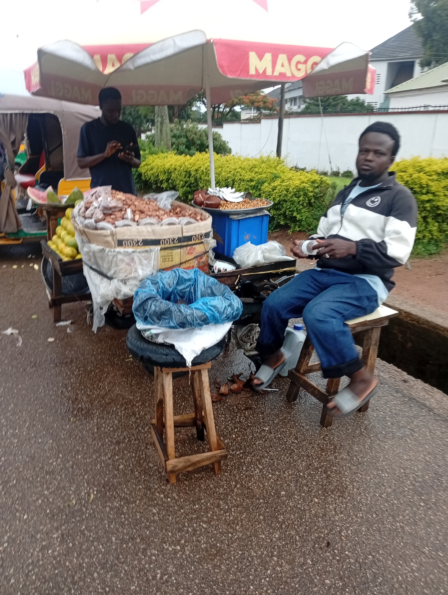 In a street scene likely in Jos, Nigeria, a young Black man is seated on a wooden stool on a paved surface, possibly a roadside. He is wearing a black and white zipped jacket over a light blue t-shirt and blue jeans, with grey flip-flops on his feet. His hands are engaged with an item, possibly a phone or a small bag. Beside him, a street vendor's stall is visible, under a large red and white umbrella with the "MAGGI" logo. The stall features a variety of goods, including what appears to be groundnuts in a large sack, lemons or limes in a tray, and possibly other snacks. A blue crate and other items are also present. The ground is wet, suggesting recent rain. In the background, a wall and lush green shrubs are visible, along with a building and trees. The overall impression is a typical street commerce setup.