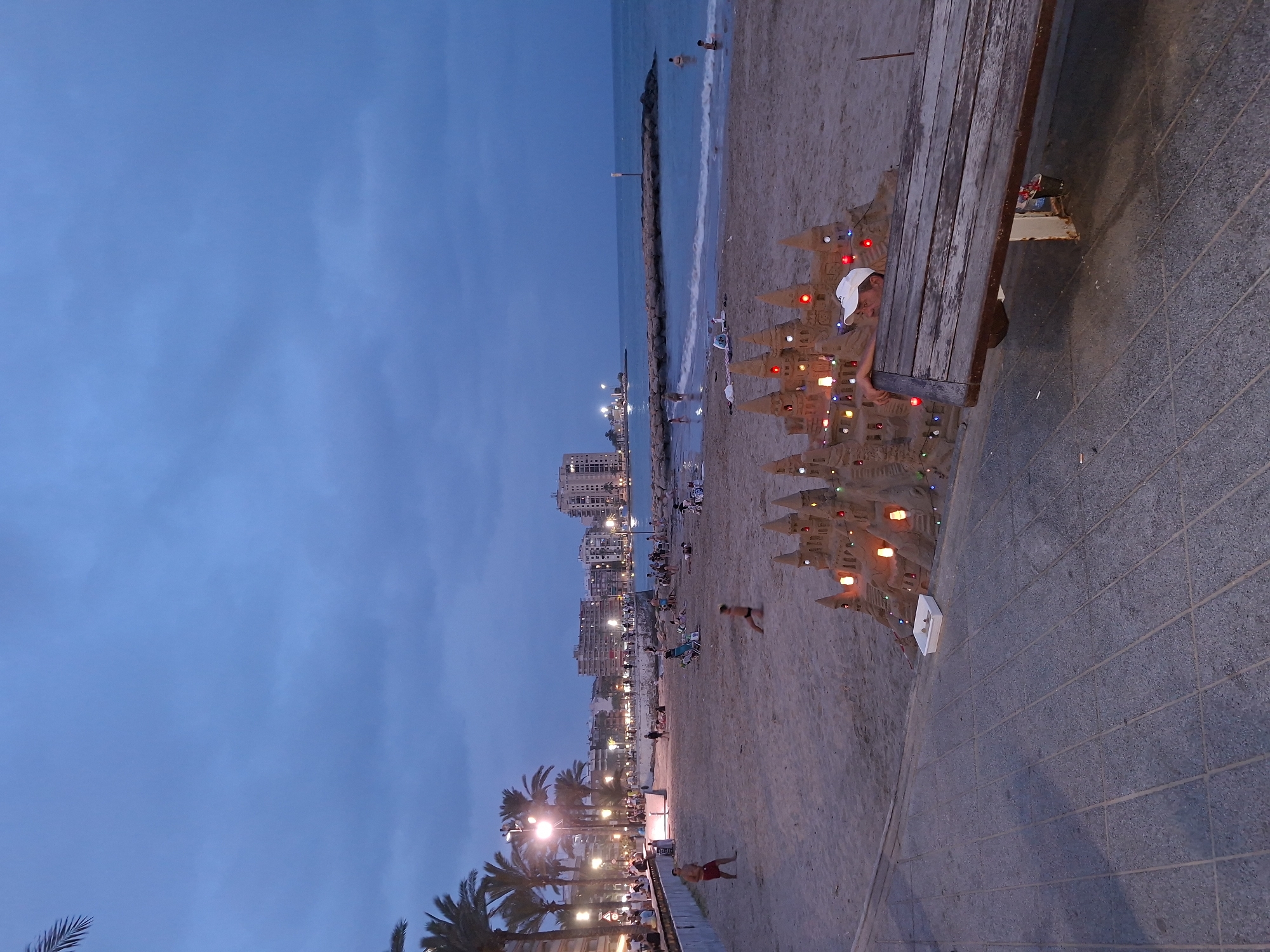 The image depicts a beach scene at dusk in Torrevieja, Spain. In the foreground, a sandcastle decorated with small lights is prominent. A wooden bench is positioned to the right, with a person wearing a white cap partially visible behind it, looking towards the sandcastle. The sandcastle sits on the sandy beach, with a white plastic tray containing some objects placed in front of it.
The beach is populated with several people enjoying the evening. Some are walking along the shore, while others are sitting or standing in small groups. In the middle ground, a section of the boardwalk or promenade is visible with palm trees and lampposts illuminating the area. Beyond the beach, the Mediterranean Sea is visible, with a rocky pier extending into the water. The background features a cityscape with buildings, suggesting an urban environment adjacent to the beach. The sky is a deep blue, characteristic of twilight.
