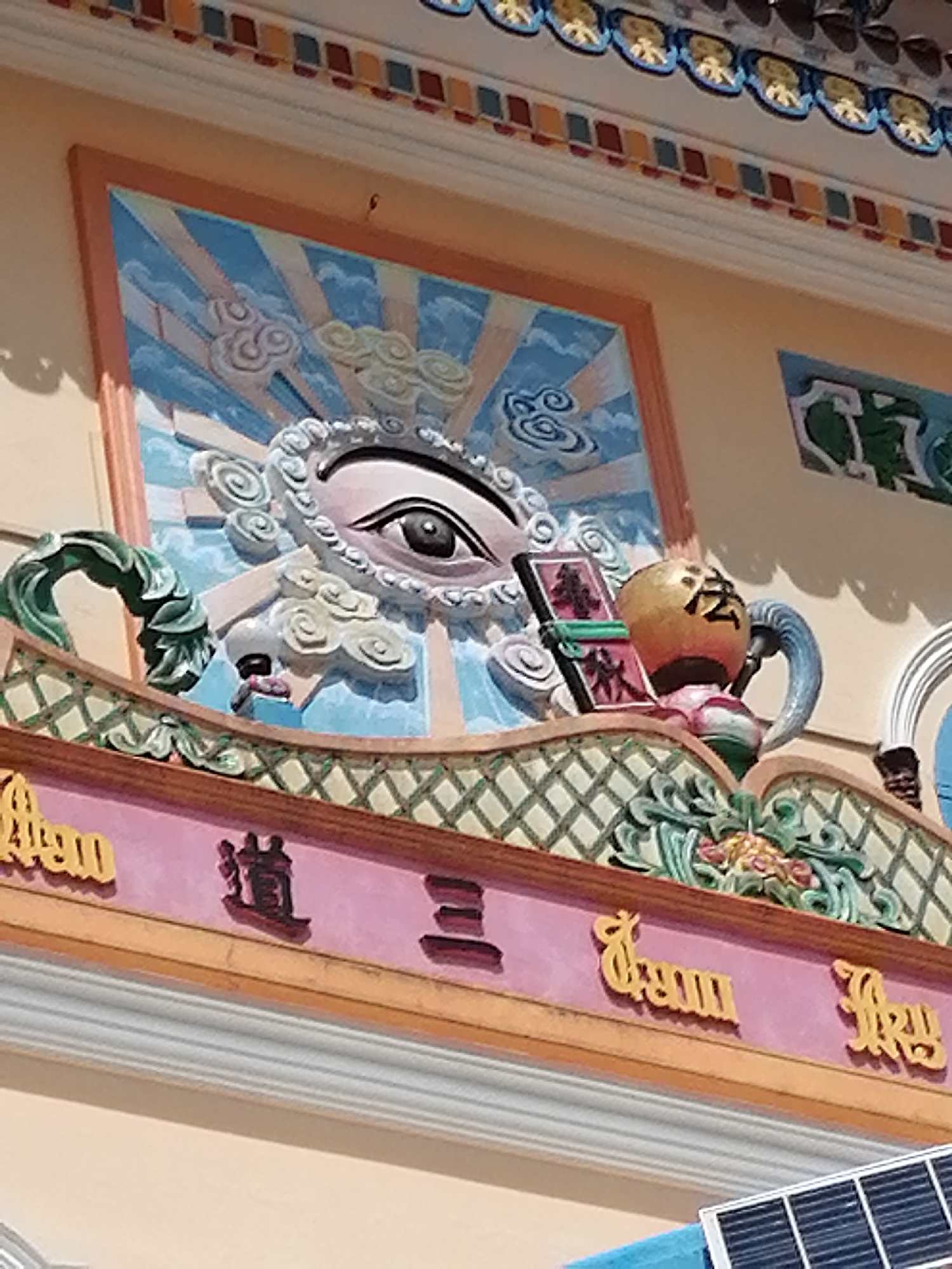 The image shows a detailed close-up of the exterior facade of a building in Lộc Quý, Vietnam. The building's architecture features intricate decorative elements, including a large, stylized eye with swirling patterns surrounding it, rendered in relief against a backdrop of blue and white rays and clouds. To the right of the eye, a golden spherical object with dark Chinese characters is visible, connected to a blue, horn-like protrusion. Below this central motif, the facade displays decorative friezes and inscriptions. A pink band runs horizontally, adorned with yellow, stylized lettering and dark, embossed characters that appear to be in Chinese or Vietnamese. The upper edge of the building has a colorful decorative border with alternating red and yellow square patterns. Green, leafy, sculpted elements are also integrated into the facade's design. The overall impression is one of ornate, possibly religious or culturally significant, architecture.