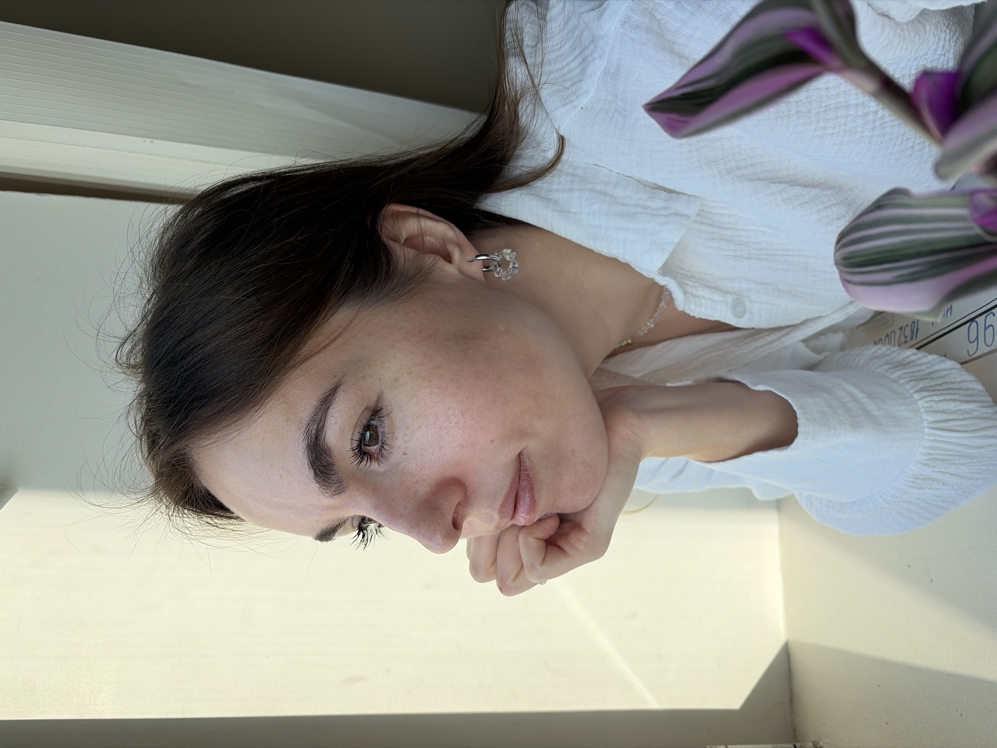 A young woman with dark brown hair and fair skin is sitting near a window. Her face is resting on her hand. She is wearing a white blouse and small, clear earrings. The background is blurred. A plant is partially visible in the foreground. There is no indication of the location being Izhevsk, Russia.