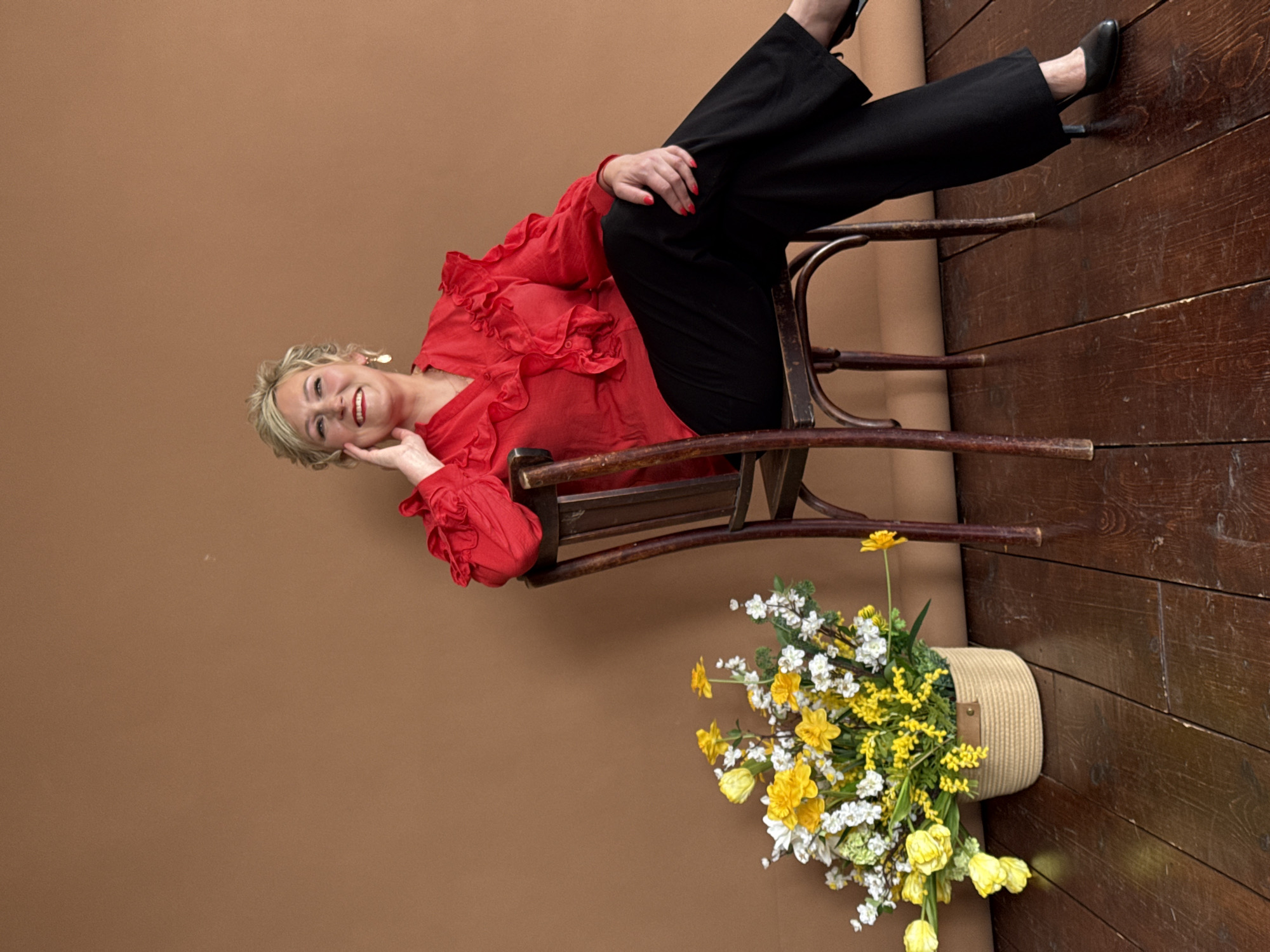 An older woman poses in a relaxed manner in an indoor setting, possibly a studio. She is reclining on her side on a dark, bentwood wooden chair, looking directly at the viewer with a warm, open smile. Her blonde hair is styled, and she is dressed in a bright red long-sleeved blouse with ruffled detailing and black wide-legged trousers, paired with black low-heeled shoes. Her left hand is gently placed on her cheek, showcasing red-painted fingernails.
The background features a plain, earthy brown wall on the left, transitioning to a dark, rustic wooden floor on the right. A woven basket filled with a cheerful arrangement of fresh or artificial flowers sits on the wooden floor in the bottom right corner. The bouquet consists predominantly of bright yellow flowers, resembling daffodils and mimosa, interspersed with small white blossoms. The overall atmosphere conveys a sense of happiness and ease. No text is visible in the image. This scene is set in what is contextualized as Chaikovsky, Russia.