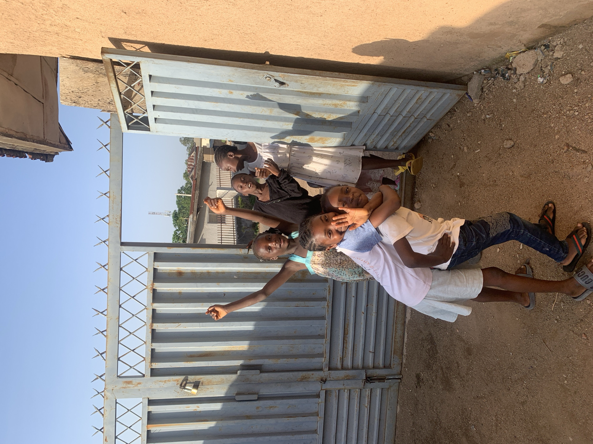 A group of five children are posing for a photograph in front of a corrugated metal gate. The gate is grey and appears to be weathered. The top of the gate has pointed spikes. The children are of varying ages and are dressed in casual clothing. Two children are in the foreground, with one boy embracing another and both looking at the camera. The other three children are standing behind them. They are smiling and appear to be in good spirits. The background shows a residential area with buildings and a clear blue sky. The scene is lit by natural sunlight, casting shadows on the ground. The overall impression is one of childhood joy and camaraderie. The image is likely taken in an outdoor setting within a city, given the presence of a gate and buildings. Without further information, it is difficult to pinpoint an exact location within Jos, Nigeria, but the scene is consistent with a typical urban residential environment in that region.