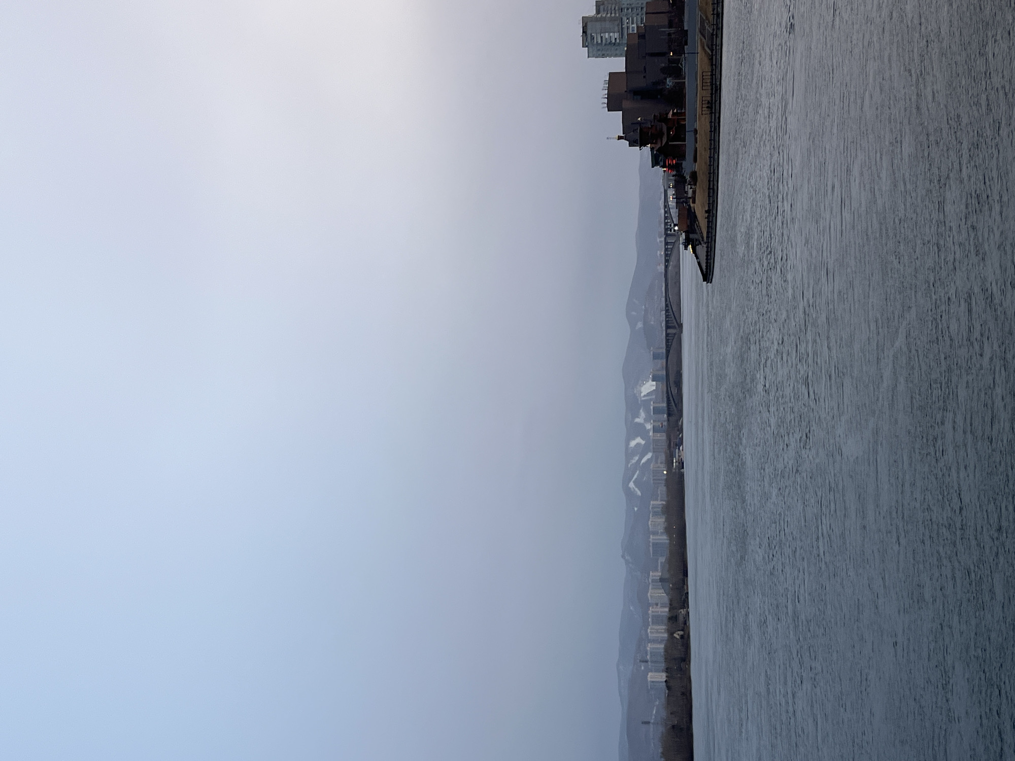 The image depicts a river landscape in Krasnoyarsk, Russia. The Yenisey River fills the right half of the frame, exhibiting a rippled surface. Adjacent to the riverbank is a line of buildings, including what appears to be a modern building complex. In the distance, mountains rise in the background. A bridge spans the river. The sky is overcast, creating a hazy gray light.