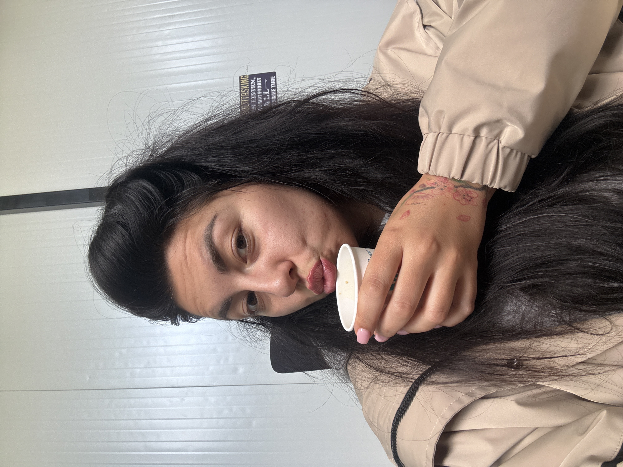 A woman is seen holding a small white cup, possibly containing a beverage, close to her lips, as if about to take a sip. She has dark hair and is wearing a light tan jacket. Her right wrist bears a tattoo. The background appears to be a white paneled wall. There are no indications of a specific location in Sofia, Bulgaria.