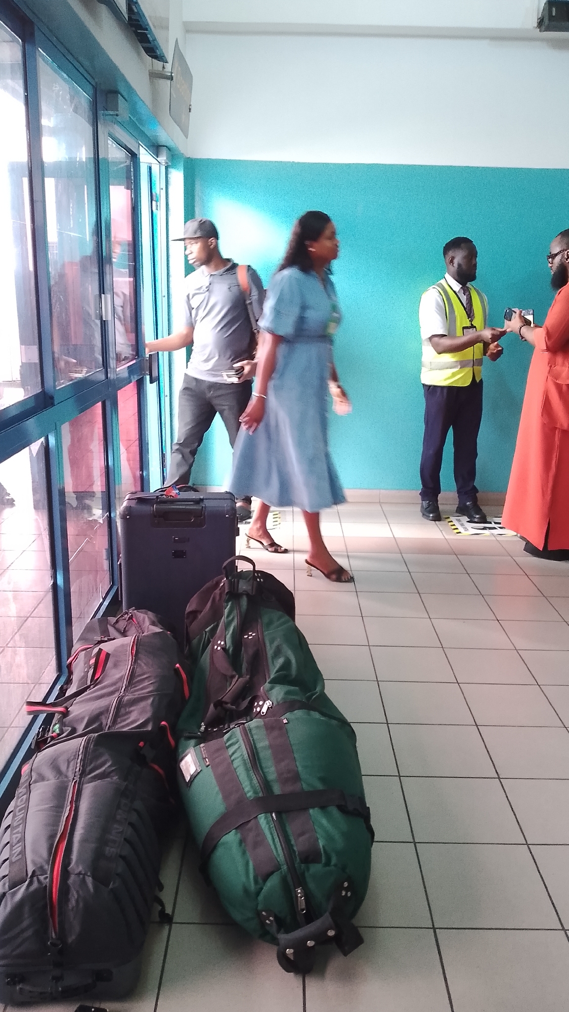 This image depicts a group of people and luggage inside what appears to be an airport terminal or a similar transportation hub. In the foreground, three large pieces of luggage are visible on a tiled floor. Two are dark gray duffel bags with red accents, and one is a large green bag with black straps. Behind these, a dark blue hard-shell suitcase stands upright.
In the middle ground, a man wearing a black baseball cap and a grey polo shirt is opening a glass door. To his right, a woman in a light blue dress is walking away from the door. Further to the right, a man in a yellow reflective vest and a dark uniform is interacting with another person dressed in orange.
The environment features a light-colored tiled floor and a turquoise wall. Large glass windows line the left side of the image, reflecting the interior and exterior. The scene suggests a moment of transition or waiting, possibly for travel. The city of Orerokpe, Nigeria, is mentioned in the prompt as the location, which would place this scene within that geographical context.