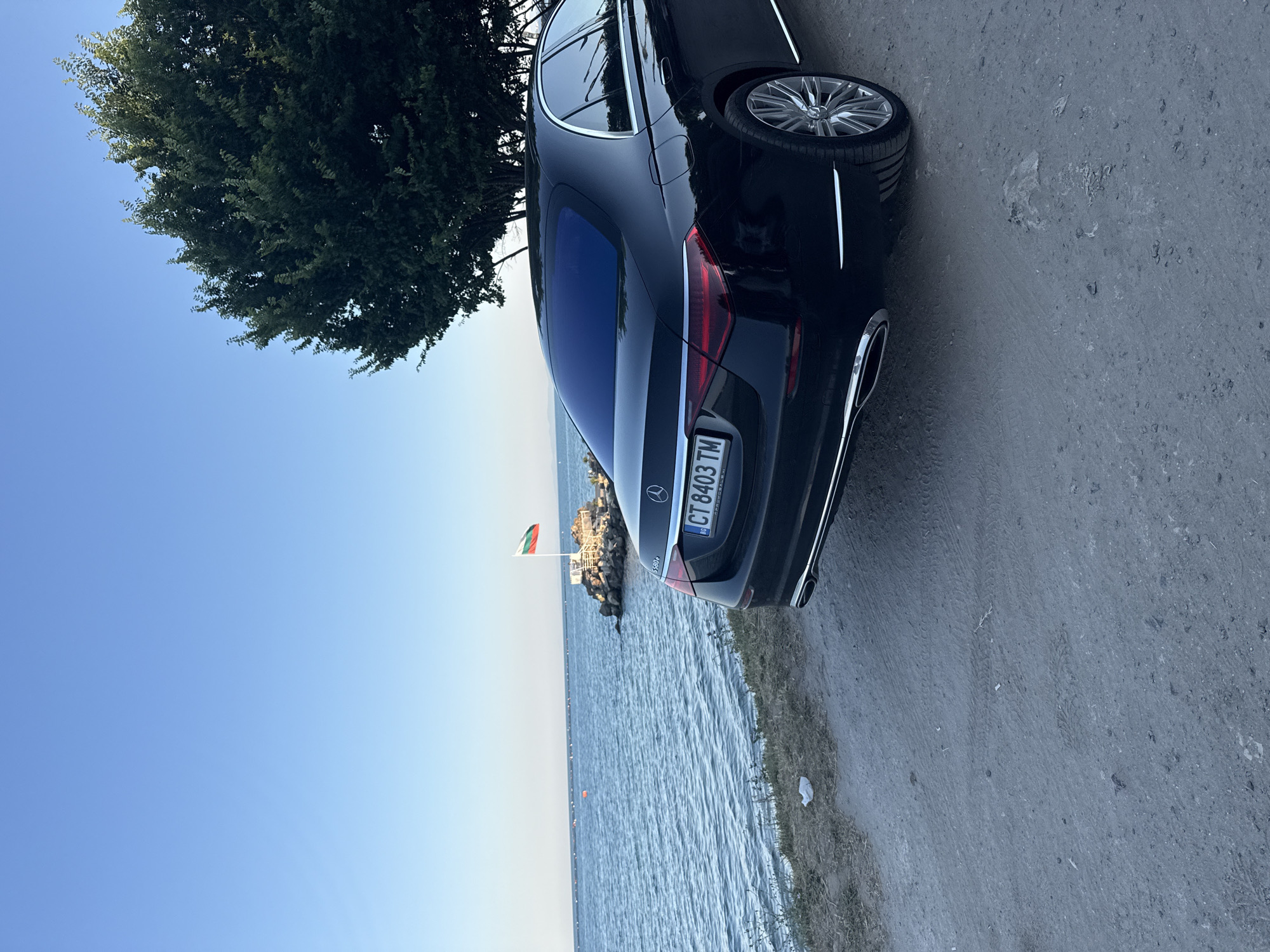 A black Mercedes-Benz sedan is parked on a paved surface near a body of water. The car's rear end is visible, showing the taillights, exhaust pipes, and license plate. The license plate reads "CT 8403 TM". A Bulgarian flag is flying on a pole in the distance, next to a rocky structure and the water. A large tree with dense foliage is situated to the left of the car, its branches reaching towards a clear blue sky. The sun appears to be low in the sky, casting a soft light on the scene, suggesting either early morning or late afternoon. The water is calm, with gentle ripples on its surface. The overall mood is serene and peaceful, with the car and the natural landscape coexisting in harmony. The image seems to be taken outdoors, possibly on a coastline or by a lake.