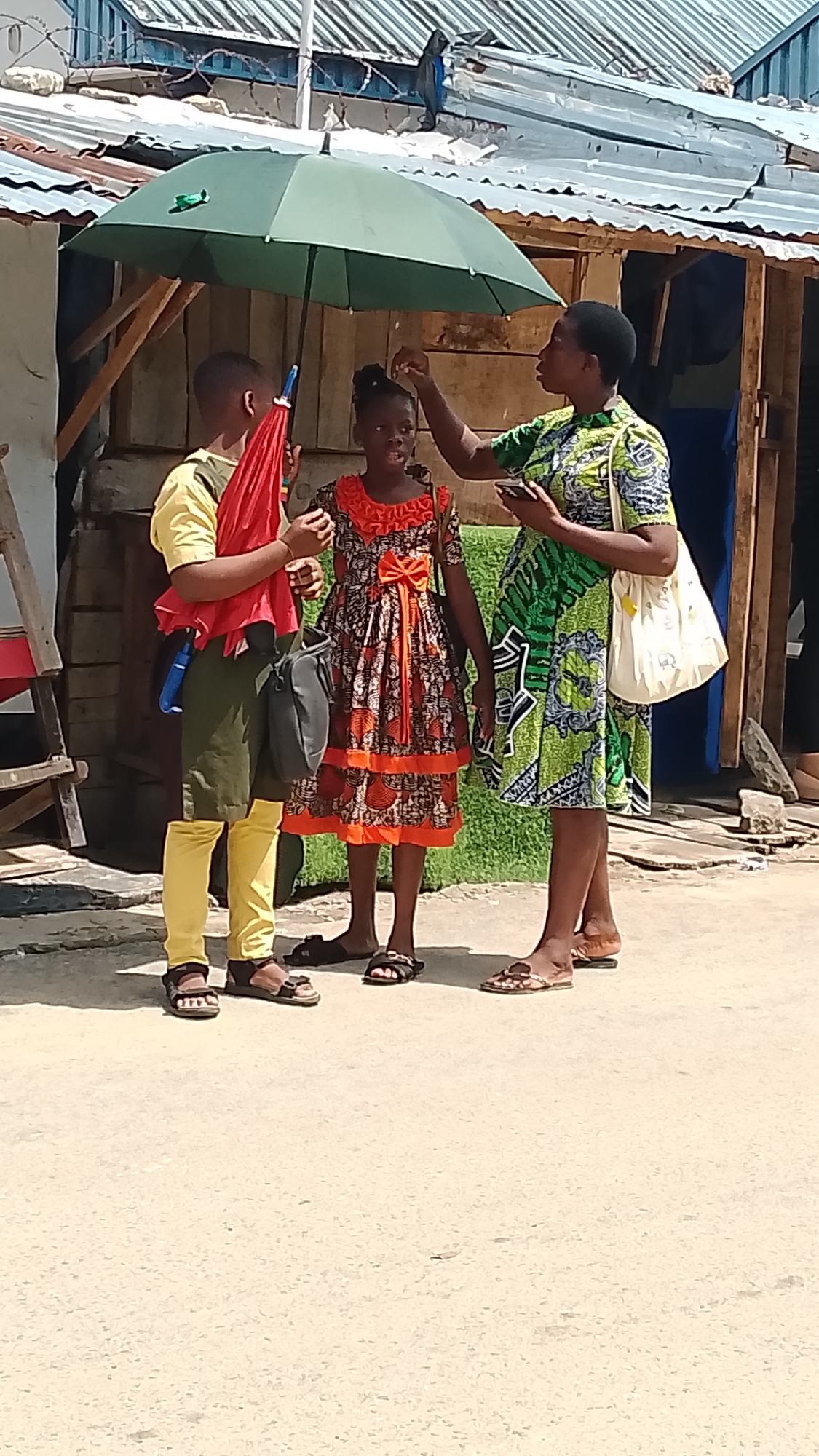 Three people stand on a sunlit paved street in front of a weathered, wooden structure, the location being the city of Unknown, Unknown. A person dressed in a yellow shirt, olive-green shorts, and yellow leggings holds a closed red umbrella. Beside them, a child wears a patterned orange and brown dress. To the right, an adult woman in a green patterned dress touches the child's head. A large green umbrella is held over them for shade. The woman also has a white tote bag over her shoulder. Each of the three individuals wears sandals. The background features corrugated metal roofing and wooden structures, suggesting a developing urban environment.