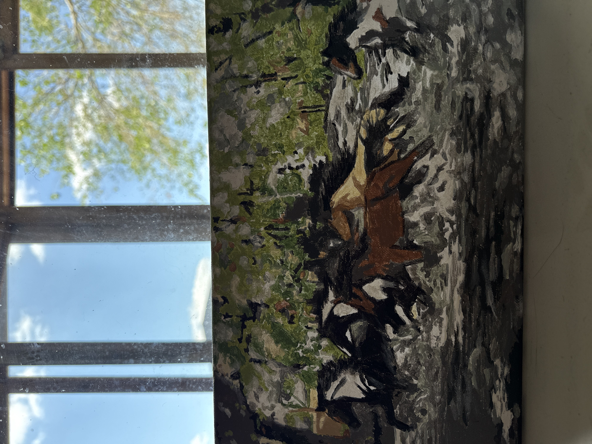 The image is a close-up, horizontal photograph of a painting depicting a herd of horses running through water, possibly a river or stream. The painting is in a somewhat abstract, impressionistic style with visible brushstrokes. The horses are rendered in various colors including brown, black, white, and tan. The background of the painting shows trees and foliage in shades of green, brown, and gray. The painting is positioned in front of a window that shows a bright blue sky with some white clouds and the blurred green leaves of trees. The window frame, with its vertical dividers, is also visible. The bottom of the image shows a plain, light-colored surface, possibly a table or shelf. There is no information within the image to suggest a location in Raivka, Ukraine.
