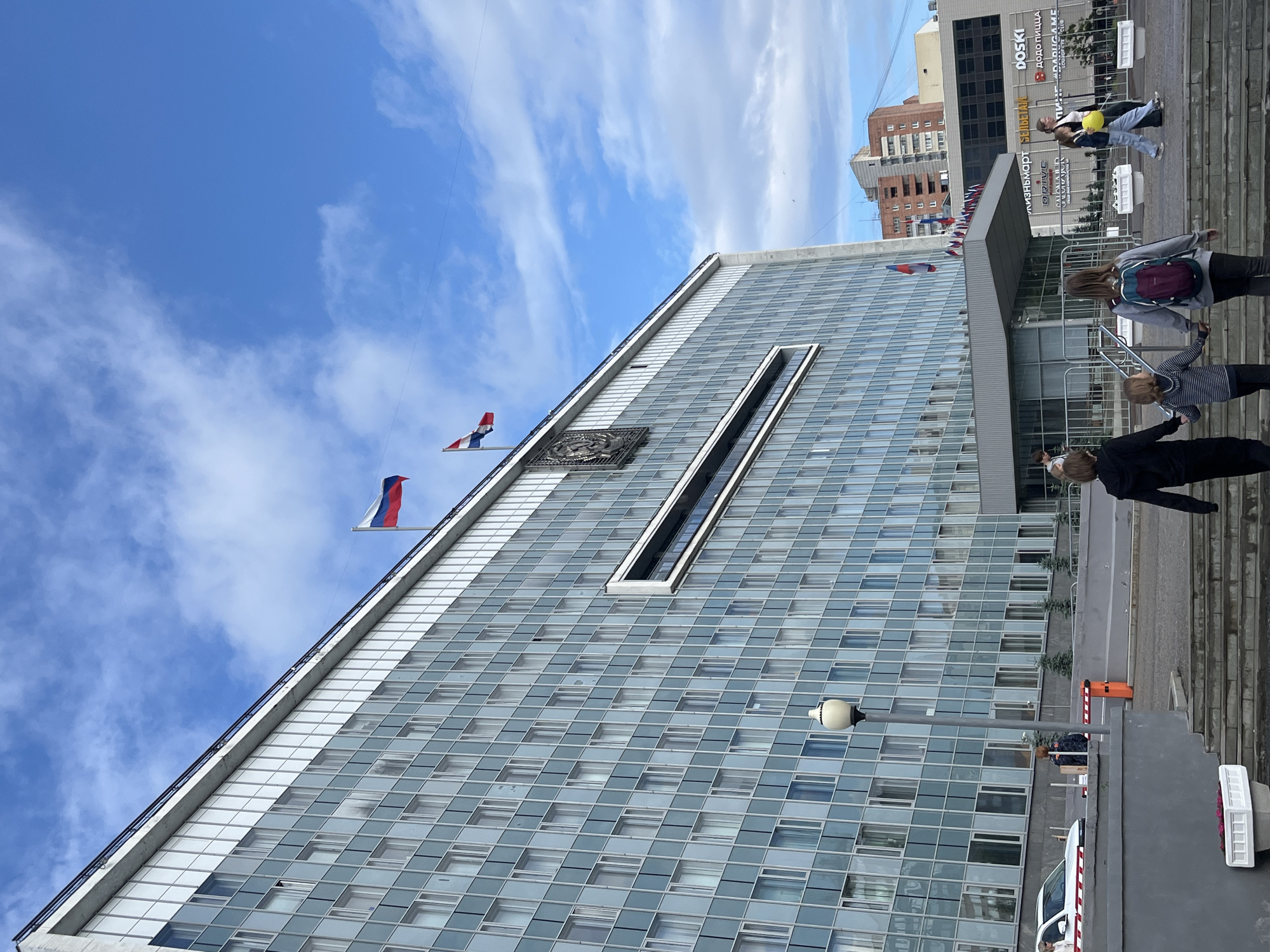 A low-angle, wide shot captures the imposing facade of a modern, multi-story administrative building, likely a government institution, in the city of Perm, Russia. The building's exterior is characterized by a grid of glass windows set in a light grey frame. Two Russian flags are prominently displayed on the building's upper level, fluttering against a bright blue sky dotted with white clouds.
In the foreground, a family group, consisting of two adults and two children, walks up a set of concrete stairs. The adults are walking hand-in-hand with the children. The adult on the left is wearing a black hooded sweatshirt and pants. The adult on the right is wearing a grey sweatshirt with a backpack and dark pants. The taller child is wearing a striped long-sleeved shirt and pants, while the smaller child is wearing a striped long-sleeved shirt and grey pants.
To the left of the building, a white car is visible, and a red and white striped barrier is partially obstructing a driveway. In the background, other buildings and structures can be seen, including a taller brick building. The scene appears to be an urban environment during daylight hours.