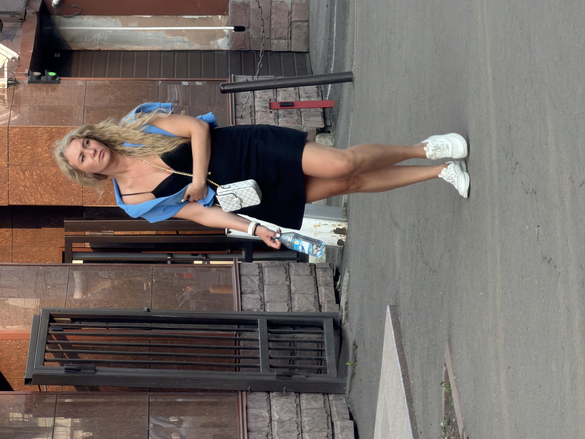 A Caucasian woman with blonde, wavy hair is walking on a paved surface, holding a bottle of water in her right hand. She is wearing a black dress, white sneakers, and a blue jacket draped over her shoulders. A small white purse with a gold chain is slung across her body. The background shows a building with a textured brown facade and a metal gate. The scene appears to be an urban environment in Krasnoyarsk, Russia, possibly near an entrance to a commercial establishment, given the building's architecture and the presence of a gate. The lighting suggests it is daytime, with sunlight casting shadows.