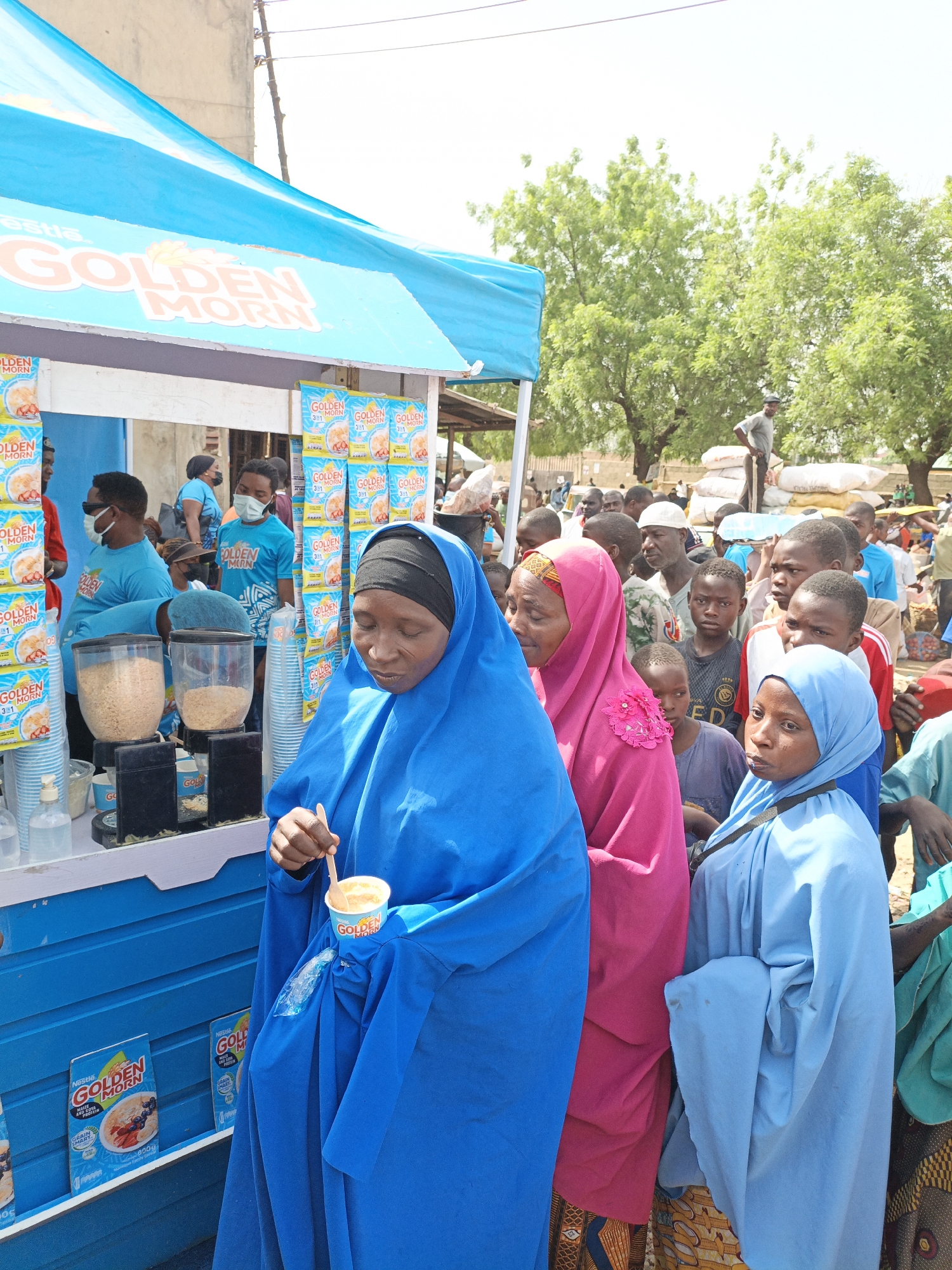 The image shows a crowd of people in Kawaji, Nigeria, gathered around a Nestle Golden Morn promotional stand. The stand is blue and white with Golden Morn branding and product displays. Three women in traditional Muslim clothing (blue, pink, and light blue hijabs) are in the foreground. The woman in blue is eating from a Golden Morn cup with a wooden spoon. Behind them, a diverse crowd of men, women, and children stand, many watching the activity at the stand. Some of the stand workers are wearing blue Golden Morn branded shirts and face masks. In the background, there are trees, buildings, and what appears to be a market or storage area with stacked sacks. The scene takes place during the day with bright sunlight.