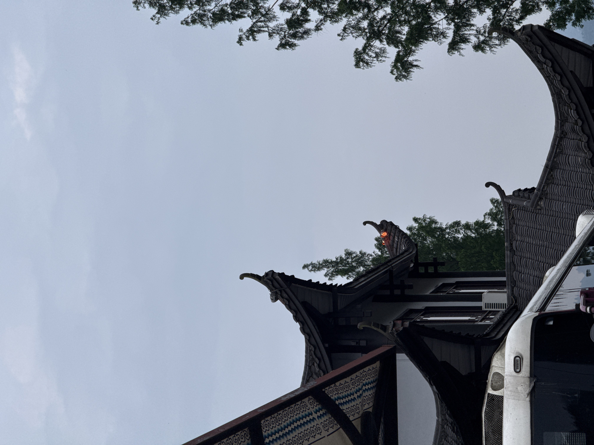 The image captures the distinctive architecture of Wulingyuan, China, with the ornate, curvilinear roofs of traditional buildings dominating the foreground. The roofs are dark, possibly tiled, and feature decorative elements, evoking an Asian architectural style. A portion of a white bus or vehicle is visible on the right, its presence suggesting the scene is in a tourist area. Trees are present in the background and upper part of the frame. The sky is overcast. The composition is tilted, giving a dynamic, low-angle perspective.
