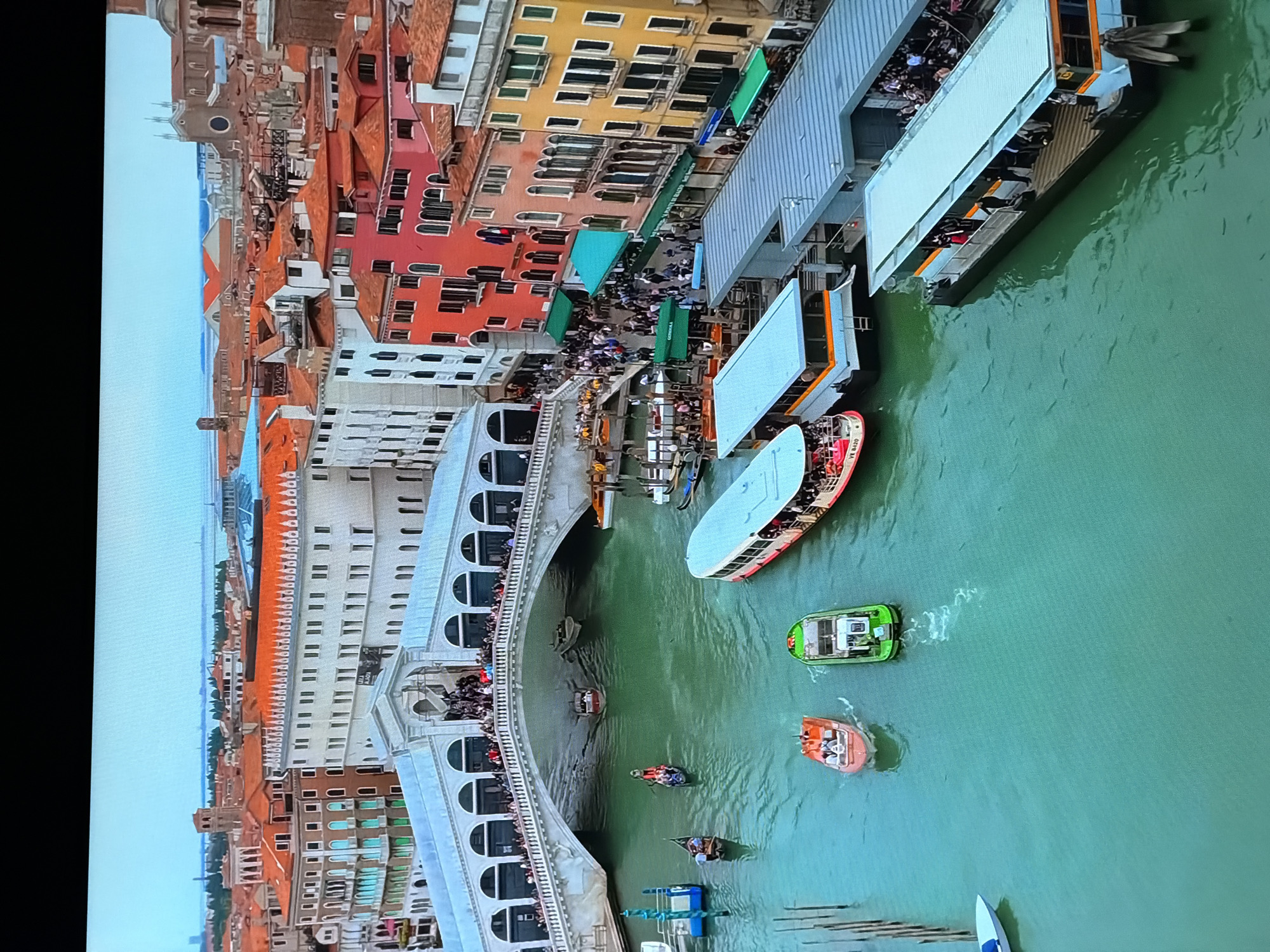 The image depicts an aerial view of the Rialto Bridge in Venice, Italy. The iconic bridge, with its white stone arches, spans a vibrant green canal. Numerous boats, including vaporettos (water buses), gondolas, and smaller motorboats, navigate the waterway, some docked at piers along the canal's edge. The canal is lined with colorful buildings, characteristic of Venetian architecture, with red, orange, and yellow facades. People are visible on the bridge, along the canal banks, and on the boats, suggesting a bustling, tourist-heavy atmosphere. The sky is clear and bright, indicating daytime. The scene captures the essence of Venice's unique urban landscape and its reliance on canals as thoroughfares. The city of Chirchik, Uzbekistan, is not depicted in this image.