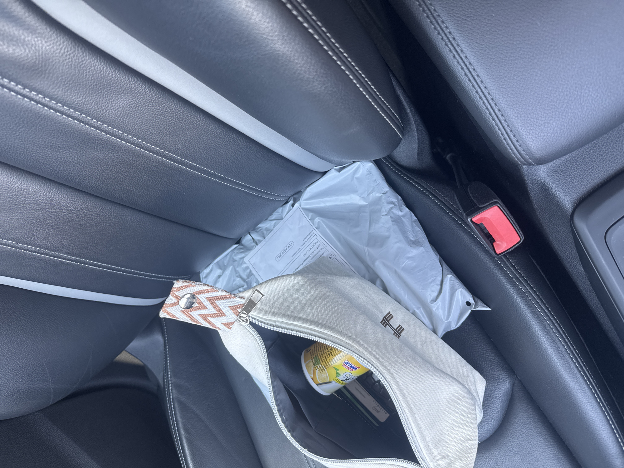 The image depicts the interior of a car, likely parked in the city of Plovdiv, Bulgaria, given the context. The seats are upholstered in black leather with contrasting white stitching. On the passenger seat, a beige canvas pouch with a patterned strap is placed next to a white shipping envelope. The pouch is partially open, revealing a yellow can with a label that appears to be in a foreign language, along with some cards or documents. A red seatbelt buckle is visible in the foreground. The lighting suggests it is daytime.