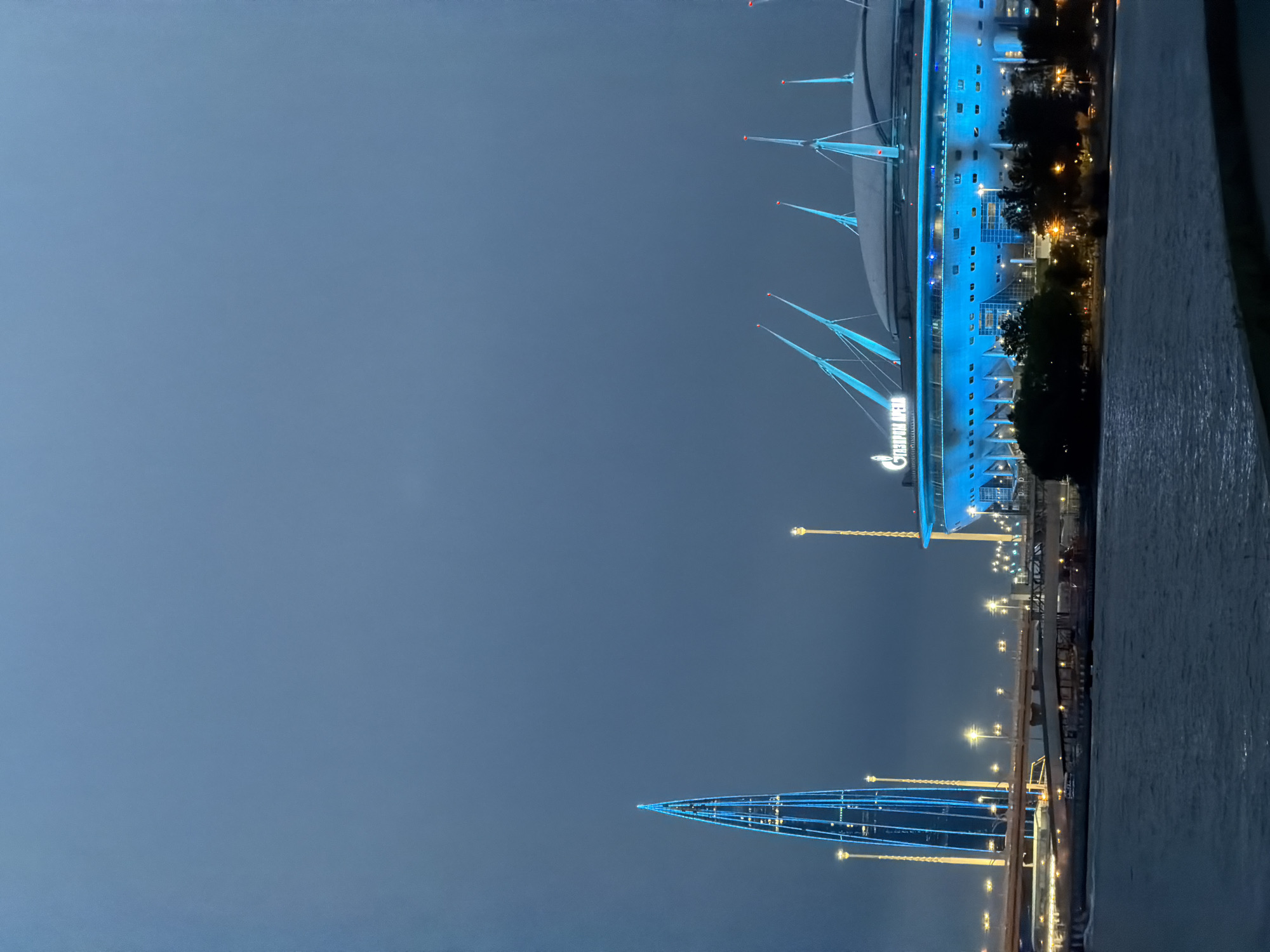 The image depicts a nighttime view of the St. Petersburg skyline, featuring the Lakhta Center skyscraper and Gazprom Arena stadium illuminated by blue and yellow lights. The Lakhta Center, a needle-shaped skyscraper, stands tall on the left side of the image, its intricate design highlighted by blue vertical lighting. To its right, Gazprom Arena, a domed stadium, is visible with its exterior glowing in bright blue light. The stadium's name, "ГАЗПРОМ АРЕНА," is illuminated in white lettering above the entrance.
A body of water, likely the Gulf of Finland or Neva River, occupies the foreground. The water is dark and reflects the lights from the surrounding structures. In the middle ground, a bridge crosses the water, with lights visible along its structure and on vehicles moving across it. Trees line the waterfront, their dark silhouettes contrasting with the illuminated buildings. The sky is a dark, hazy blue, characteristic of an evening or night scene, with clouds obscuring the stars. No people are visible in the image.