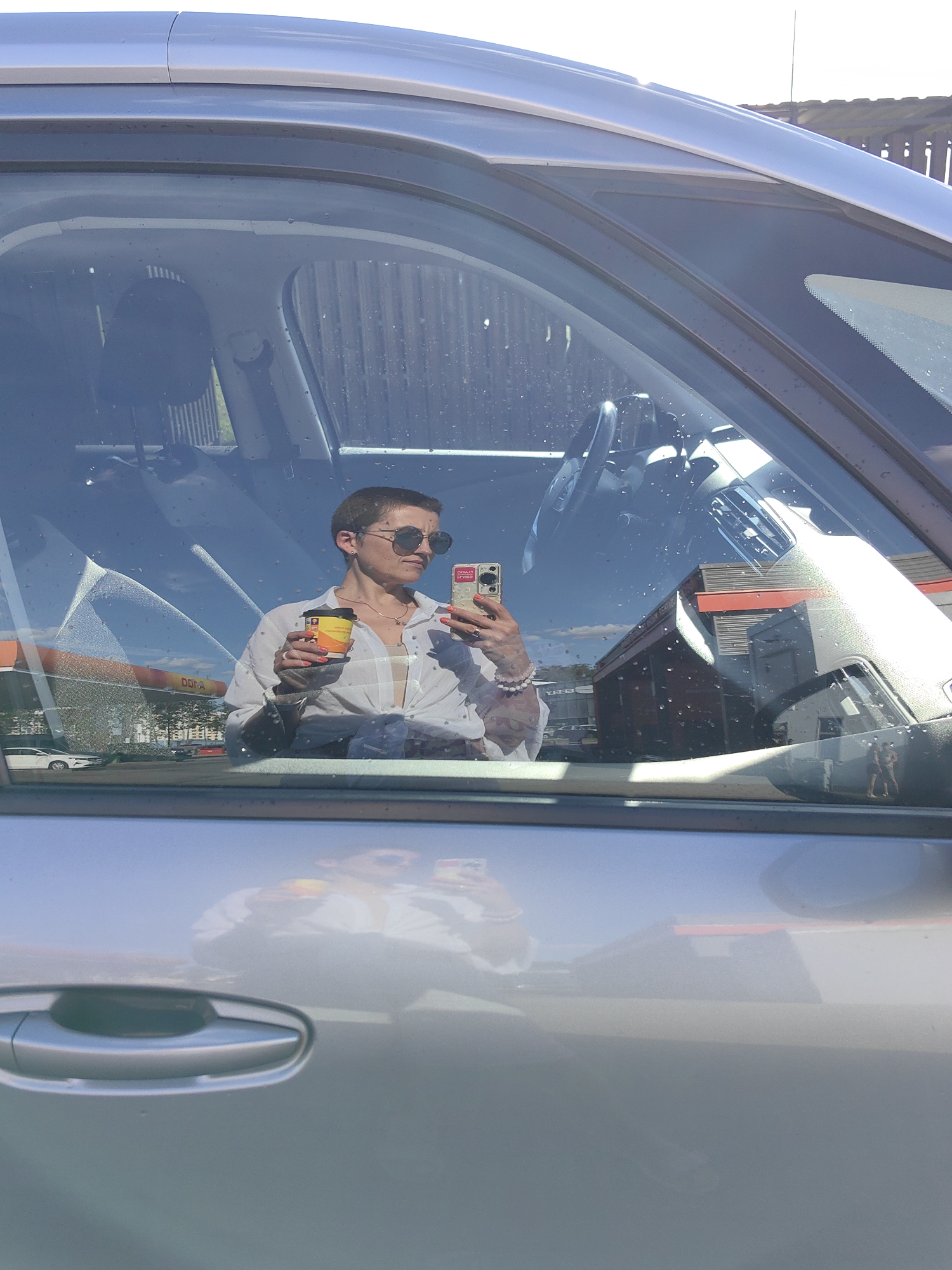 The image is a mirror selfie taken from inside a car, reflecting a woman in the driver's seat. She has short, dark hair, wears sunglasses, and a white button-down shirt tied at the waist, revealing a tan camisole underneath. She is holding a coffee cup in her left hand and a smartphone in her right, with which she is taking the photo. She wears a necklace and a beaded bracelet on her right wrist, and her nails are painted orange. The reflection shows the interior of the car, including the steering wheel and dashboard.
Outside the car, visible through the windshield, is a gas station with orange and white signage. The sky is blue with some clouds, suggesting it's daytime, likely sunny. The scene appears to be in an outdoor setting, possibly a parking lot or road near the gas station. The overall mood conveyed by the image is casual and perhaps a moment of pause or relaxation during a day.
There is text on the coffee cup and the phone case. The coffee cup has branding that includes "DOUWE EGBERTS". The phone case appears to have some text or graphics on it, but it is not clearly legible. The gas station sign also has text, but it's too blurry to read.
The provided location context of Шчомысліцкі сельскі Савет, Belarus, helps place the scene geographically.