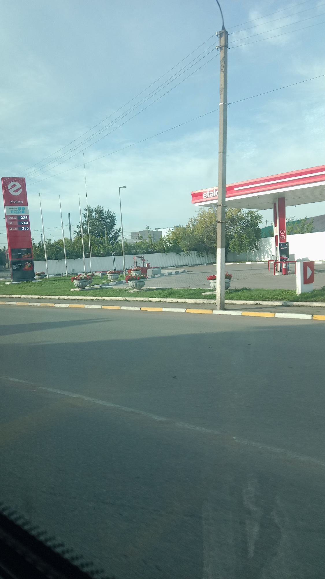 The image shows a gas station in Petropavl, Kazakhstan. The gas station is branded "etalon". A red and white canopy covers the gas pumps. There are planters with red flowers in front of the gas station. A sign on the left side of the image displays fuel prices for 92, Diesel, and 95 octane gasoline. The sky is blue with some clouds. A street and a sidewalk are visible in the foreground.