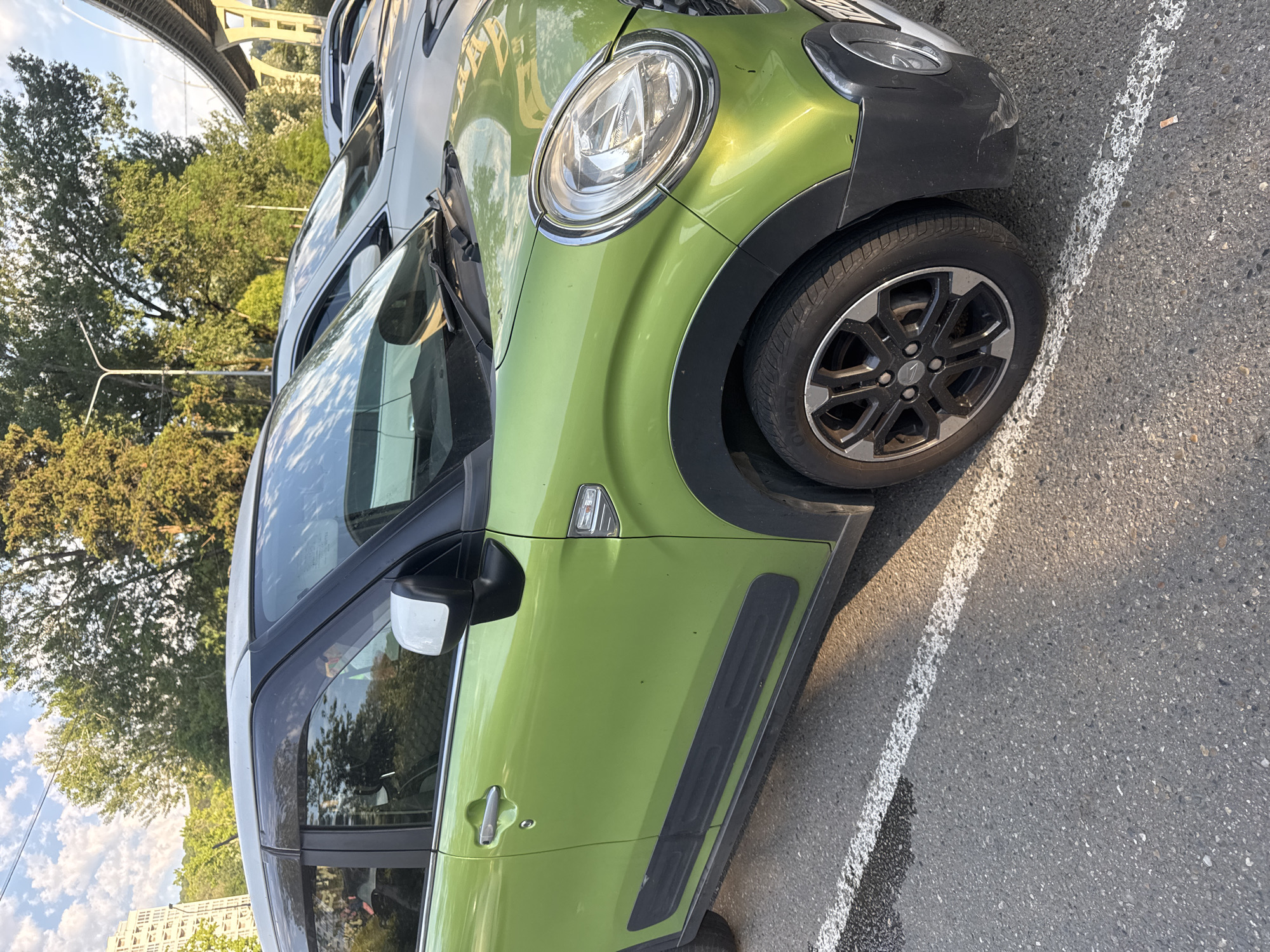 A green car is parked in a parking lot. The car is a small hatchback with a white roof. It has a black trim around the wheel wells and along the bottom of the doors. The car is parked next to other cars, and there is a white line painted on the asphalt to mark the parking space. In the background, there are trees and a building. There is also a bridge in the distance. The scene appears to be a sunny day.