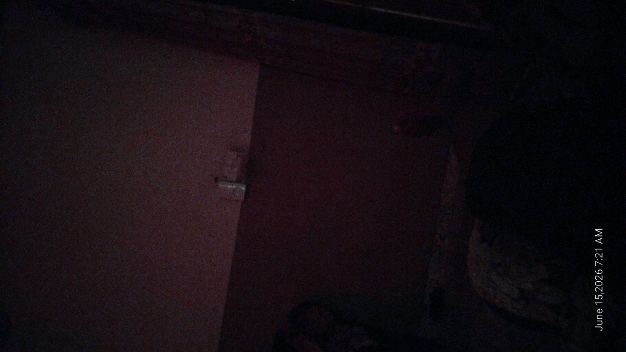 The image shows a dimly lit interior, possibly a bedroom. The lighting suggests it is either early morning or late evening, which is consistent with the timestamp "June 15, 2026 7:21 AM." The walls are a muted brown color. On the wall, near the top center of the frame, there appear to be a few small objects, possibly light switches or outlets. In the foreground, a large dark shape dominates the lower half of the image, suggesting a person lying down or sleeping. To the left of this shape, a patterned fabric is visible, possibly part of bedding or clothing. The overall scene is dark and indistinct, with limited detail visible due to the low light conditions. No specific landmarks or features that would indicate the location in Howayi, Nigeria, are discernible.