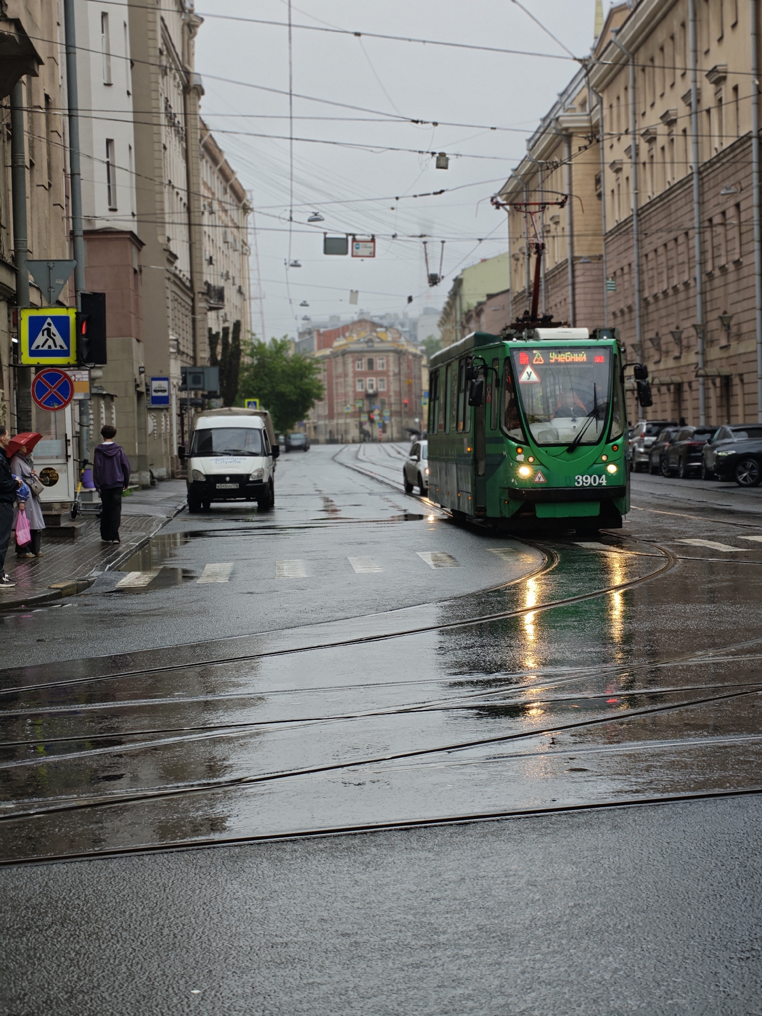 A green tram with the number 3904 is driving on a wet street in front of a building. The tram has a sign that reads "УЧЕБНЫЙ" (Uchebnyy) which means "training" in Russian. There are other cars and a white van on the street. There are people standing on the sidewalk under umbrellas, one person wearing a purple hoodie. There are traffic lights and street signs visible. The street is wet and reflects the lights of the tram, indicating it has recently rained or is raining. The sky is overcast, suggesting a cloudy or rainy day. The buildings on either side of the street are typical of an urban environment, with multiple stories and windows. The tram tracks are visible on the wet asphalt. The scene appears to be a street in a city, possibly in Russia, given the text on the tram. The overall mood is subdued due to the weather conditions.