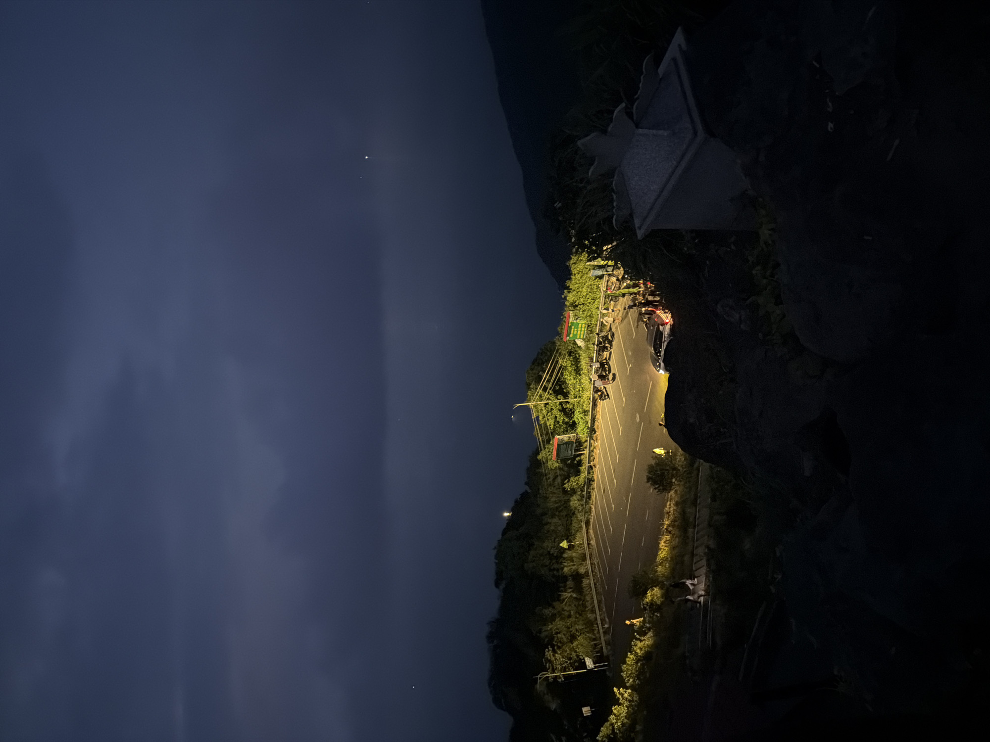 The image shows an outdoor scene at night, likely a parking area on a hillside. The sky is dark blue with a few visible stars. A paved parking lot is brightly lit by streetlights, revealing parked cars and motorcycles. A roadside fence separates the parking lot from a dense area of trees and vegetation. The foreground includes dark, rocky terrain, partially obscuring the view of the parking area. A structure resembling a monument or architectural feature is situated on the rocky terrain. The city of Đà Nẵng, Vietnam, cannot be definitively confirmed as the location.