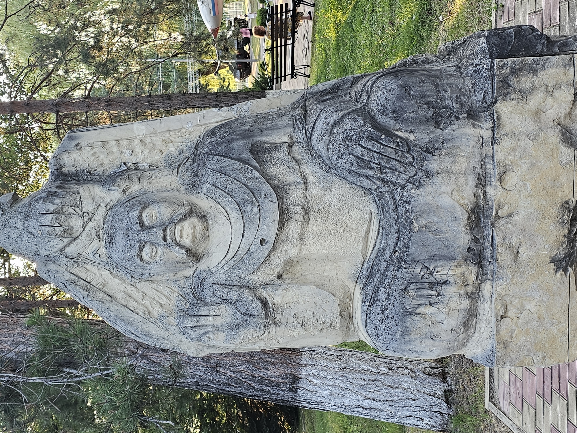 A large, weathered stone sculpture of a seated figure with a mustache is centrally located in a park-like setting. The sculpture depicts a person with a stern expression, wearing draped clothing. The figure's right hand is raised with fingers extended, while the left hand rests on its lap. The sculpture is textured and appears to be carved from a single piece of stone.
In the background, several trees with rough bark are visible, along with a grassy area. Further back, a park bench is occupied by a person sitting and facing away from the camera. Beyond the bench, military vehicles and a small aircraft are present, suggesting the location might be an outdoor museum or memorial park. The ground in front of the sculpture is paved with interlocking bricks in a red and tan pattern. The sky is partially visible and appears to be a bright, clear day.
This sculpture is located in the city of Cherkessk, Russia, specifically at the Victory Park (Park Pobedy).