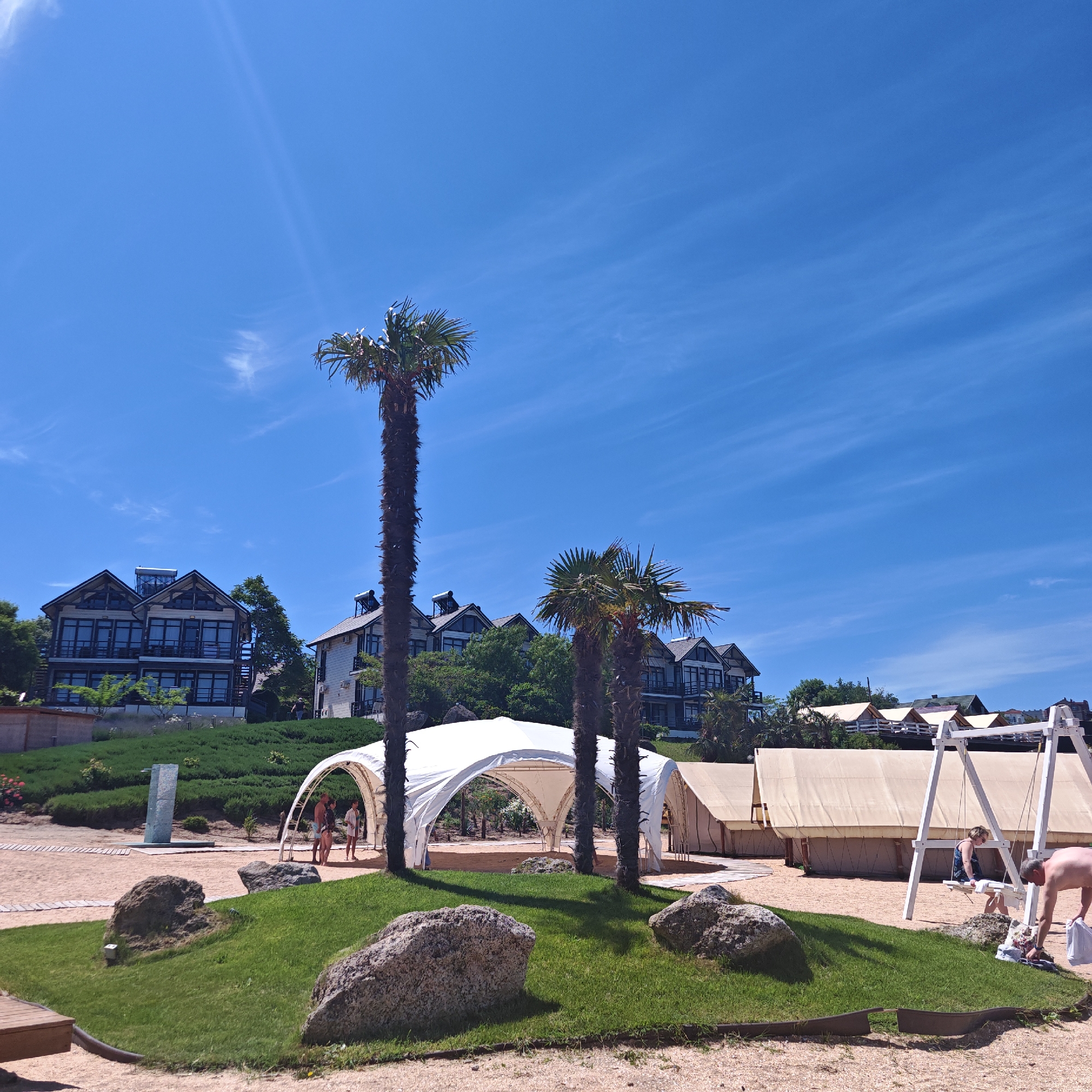 This image captures a bright, sunny resort or beach setting under a clear blue sky with faint cirrus clouds and visible sun rays. In the background, several modern multi-story buildings with dark-framed windows and balconies are situated on a lush green hillside, some featuring solar water heaters on their roofs. The mid-ground is dominated by a sandy area, where a large white arched tensile structure (tent or canopy) stands prominently between two tall palm trees. To the right, a series of light-colored rectangular canvas tents, resembling glamping accommodations, are arranged along a wooden pathway. A white wooden swing set is positioned on the sand near these tents. The foreground consists of sandy ground interspersed with patches of green grass and large granite boulders. A tall, rectangular modern water feature or sculpture stands on the left side of the sandy expanse. Several individuals are present: three people are visible beneath the white tensile structure, while near the swing set, a woman sits on the swing and a shirtless man bends over the sand, appearing to handle towels or clothing. The scene is set in an unspecified location within the city of Unknown, Unknown.