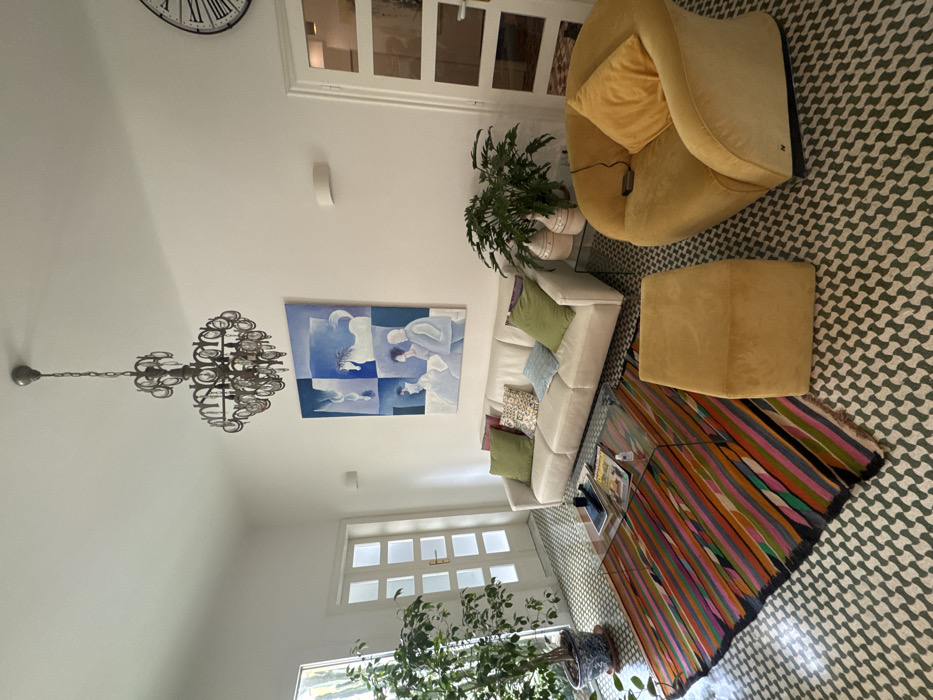 This image displays a brightly lit interior living space located in Lecce, Italy. The room features white walls and a floor covered in light green and white geometric-patterned tiles. A multi-tiered, dark metal chandelier with numerous small reflective elements hangs from the white ceiling.
Centered on the wall above a white three-seater sofa is a large rectangular painting, primarily in shades of blue and white, depicting abstract figures and a horse across four distinct panels. The sofa is accessorized with five throw pillows: two solid olive green, two with intricate patterns, and one light blue.
In front of the sofa, a transparent glass coffee table, with magazines and a small white object on its surface, sits atop a rectangular rug. This rug displays a pattern of diagonal stripes in multiple colors including various shades of red, orange, pink, green, and black. To the right of the rug, a mustard yellow velvet armchair with a matching pillow faces the sofa. Adjacent to the armchair, a rectangular mustard yellow velvet ottoman is positioned.
On the left side of the room, a tall green plant in a blue and white patterned pot stands near a white door composed of multiple glass panes. A window is partially visible behind this plant. On the right side, a smaller green plant in a cream-colored pot rests on a glass-topped console table, flanked by two large, cream-colored decorative vases. A white door with frosted glass panels is partially visible on the far right, next to a large circular wall clock featuring Roman numerals. The room is unoccupied, and no active interactions or actions are depicted.
