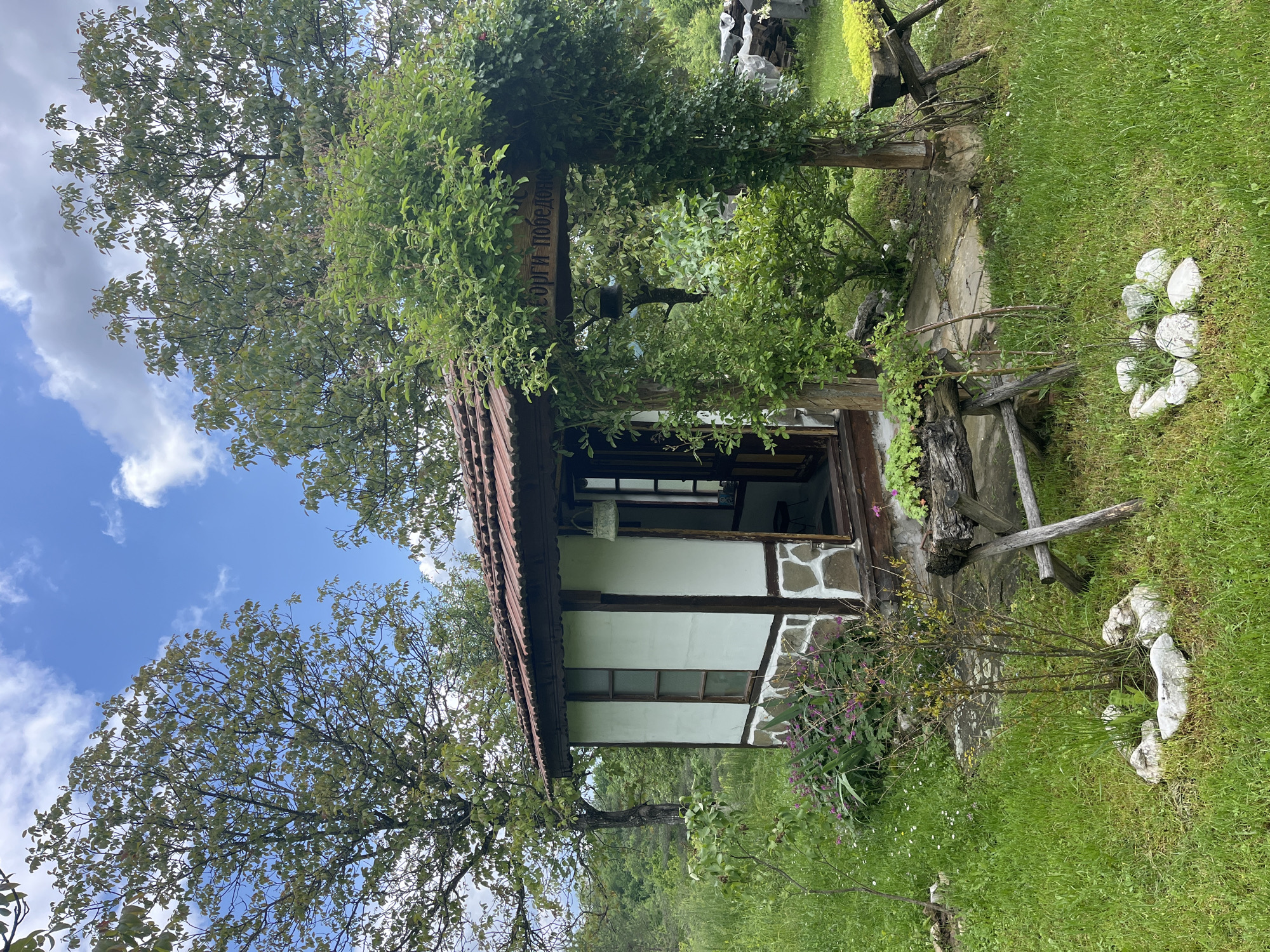 The image shows a quaint, one-story building located in the village of Shopite, Bulgaria. The structure has a steeply pitched, reddish-brown tiled roof and walls that are partially white stucco and partially stone. Brown wooden beams frame the windows and doors. A wooden sign, covered in foliage, is mounted on a post near the entrance. Lush green grass surrounds the building, and a scattering of white stones forms a border along the edge. Trees with leafy branches frame the building against a blue sky dotted with white clouds.