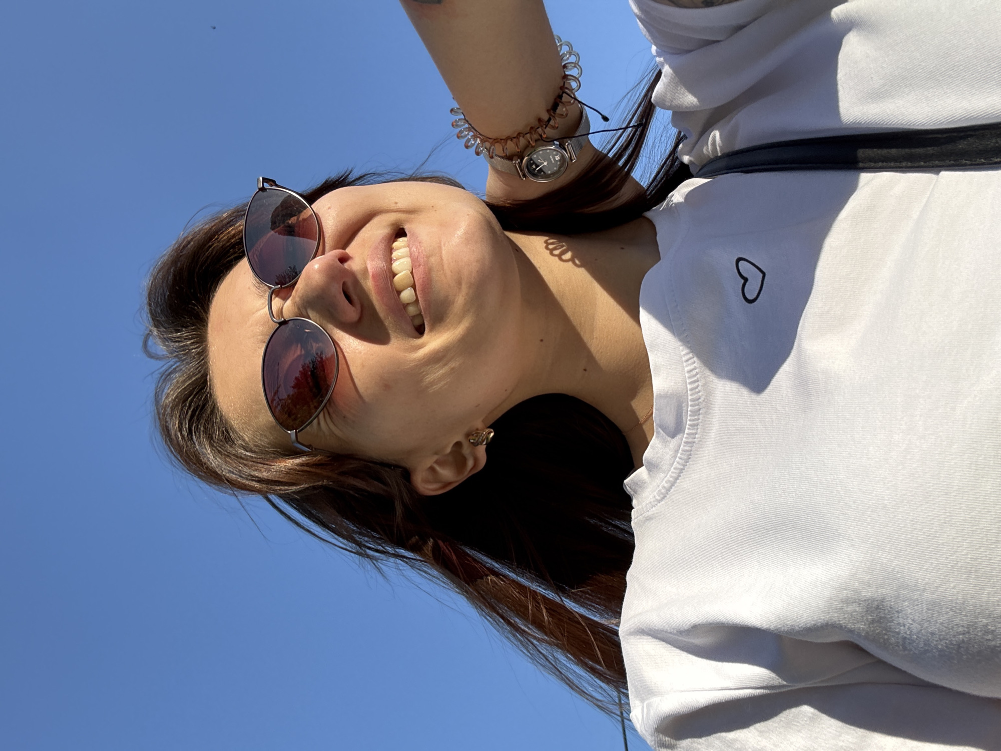 The media file is a close-up, low-angle selfie of a young woman against a bright blue sky. The woman is wearing sunglasses with red-tinted lenses and a white t-shirt that has a small black heart outline embroidered on the left chest. She is smiling broadly, her mouth open to reveal her teeth. Her hair is dark brown and appears windblown. She is wearing a silver watch on her left wrist, which is adorned with a coiled bracelet. A dark strap, possibly from a bag, is visible across her t-shirt. The sun appears to be shining brightly, suggesting it is daytime. The clear blue sky indicates good weather. The image was taken in Novokuznetsk, Russia.