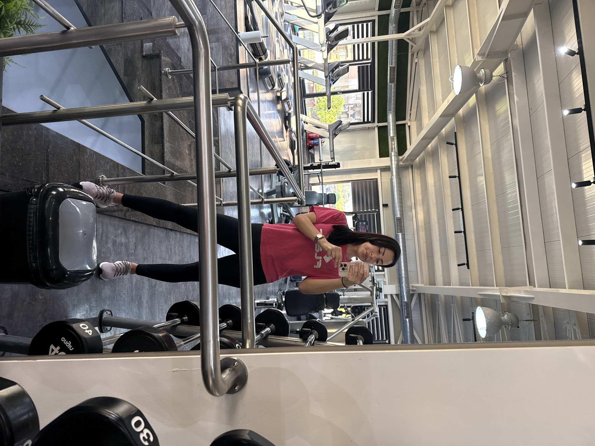 The image captures a woman in a gym setting, likely taking a selfie. She is dressed in a pink t-shirt and black leggings, holding a cell phone in her hand. The background reveals a modern gym environment with various exercise machines and equipment. Weights are visible in the foreground, along with a mirror reflecting the woman and the gym's interior. The architecture suggests a well-lit, structured space with white beams and supporting structures. The location appears to be in an urban environment, judging by the view through the gym windows.