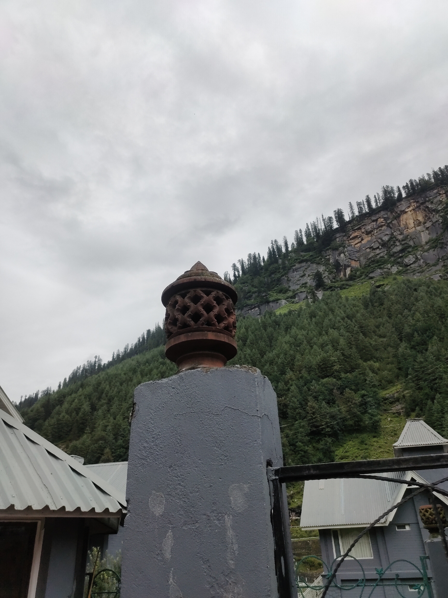 The image depicts a cloudy sky over a mountainous region with dense evergreen trees covering a steep slope. In the foreground, a weathered, reddish-brown ornamental post with intricate carvings is visible, topped by a decorative finial. This post stands on a grey, textured pillar, which is partially obscured by a black metal fence. To the left and right of the pillar, parts of buildings with corrugated metal roofs are visible, suggesting a residential or commercial area nestled within the landscape. The overall scene captures the atmosphere of Burwa, India, a location known for its mountainous terrain and traditional architecture.