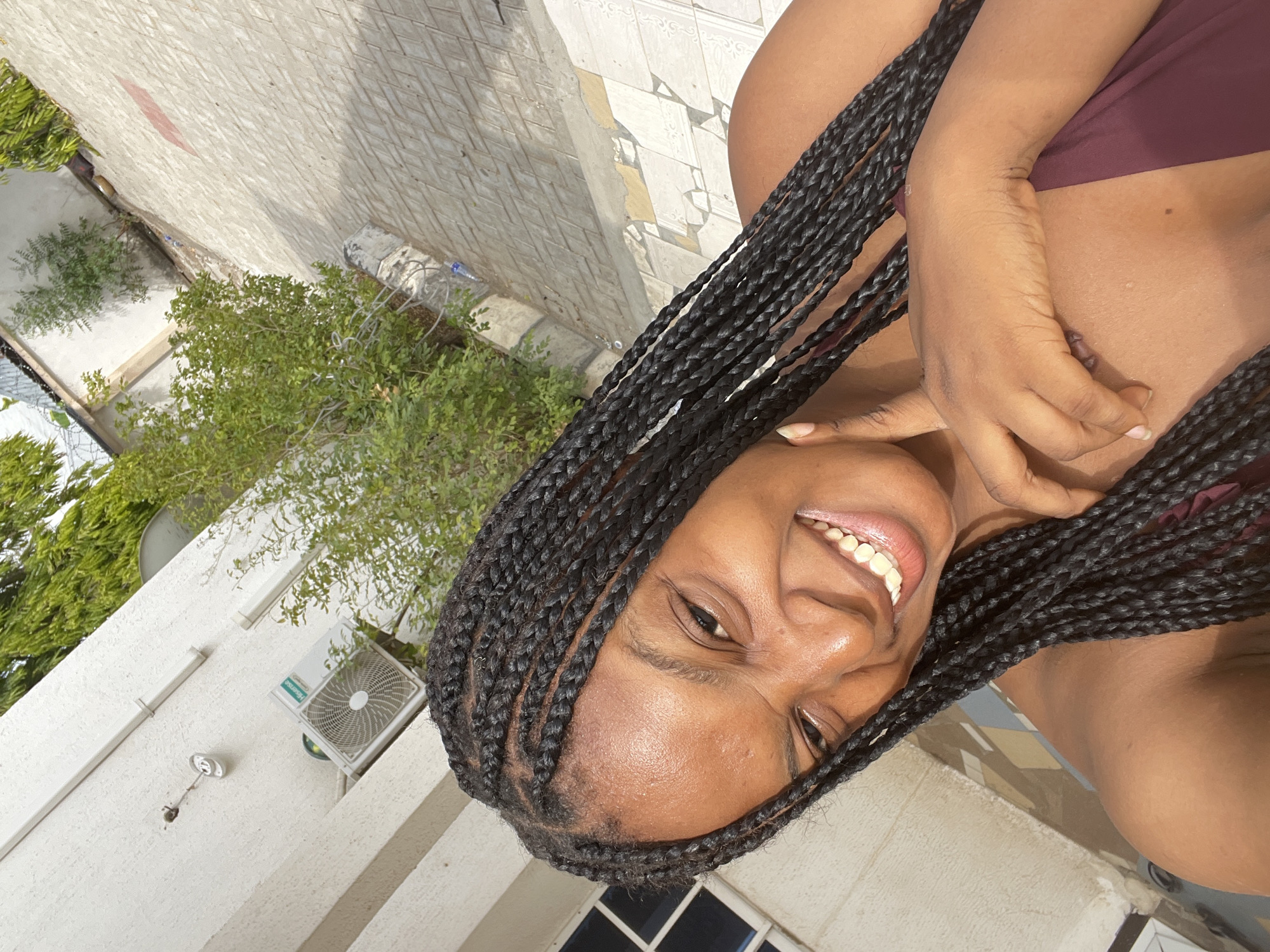 A human subject with a dark complexion and long, black box braids occupies the foreground, positioned centrally and looking directly at the viewer. The subject's mouth is open in a wide smile, revealing upper and lower teeth. The right hand is raised, with the index finger making contact with the right cheek near the mouth. The subject wears a dark, possibly maroon, garment exposing the shoulders and upper chest. The background features a white, textured building facade with a mounted Hisense outdoor air conditioning unit, white conduits, and a rectangular window with dark panes. Green foliage, including shrubs and trees, is present around the building. The ground is paved with diamond-patterned light-colored tiles, with a segment of broken white and grey tiles, and a blue plastic bottle visible near a concrete structure. The scene is illuminated by strong overhead sunlight. This location is Lokoja, Nigeria.
