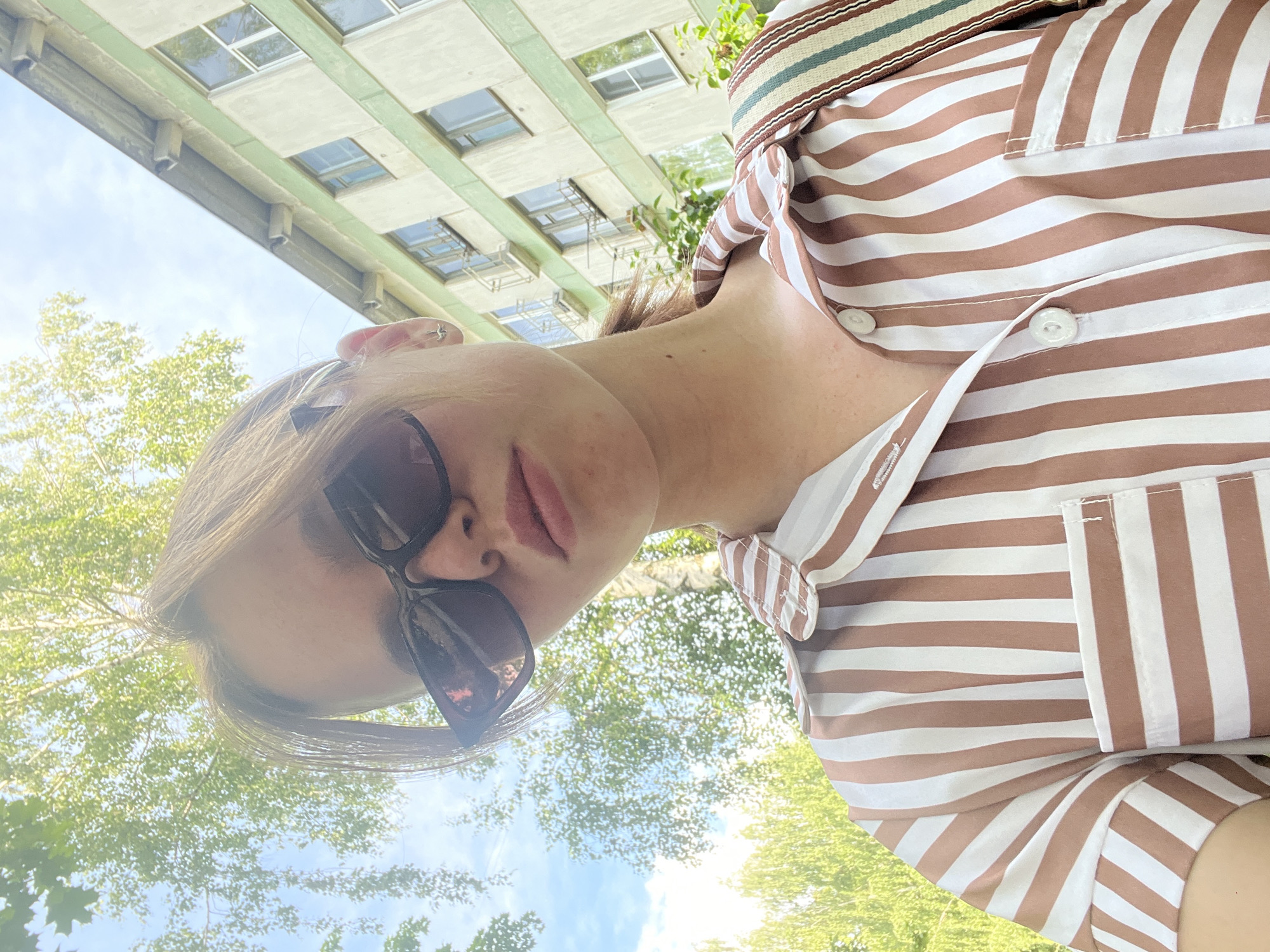 A young woman with light brown hair, wearing large dark sunglasses and a brown and white striped button-up shirt, takes a selfie outdoors. The background shows a pale blue sky with scattered clouds, leafy green trees, and a light-colored building with green trim and multiple windows. The woman has a neutral expression and is looking directly at the camera. A portion of a striped strap is visible over her right shoulder. The scene appears to be in a residential area of Chaikovsky, Russia.