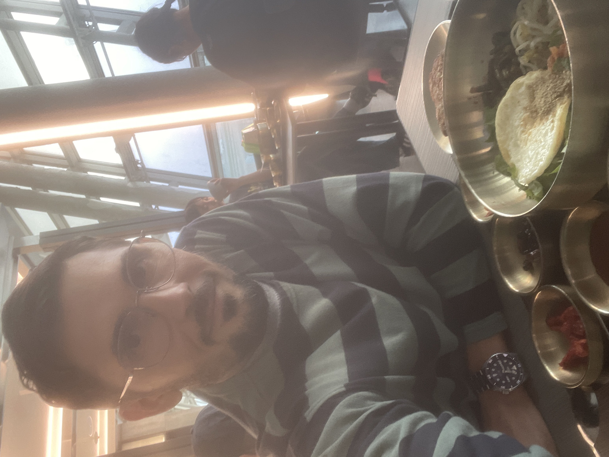 The image is a close-up, slightly blurry selfie of a man wearing glasses and a horizontally striped long-sleeved shirt. He is seated at a table, likely in a restaurant, with other diners in the background. In front of him is a metal bowl containing a dish that appears to be bibimbap, with rice, vegetables, and a fried egg. There are also small bowls with red and dark contents, possibly side dishes or sauces. The background shows a large window with vertical lighting elements, suggesting an indoor setting with a view. This scene is taking place in Incheon, South Korea.