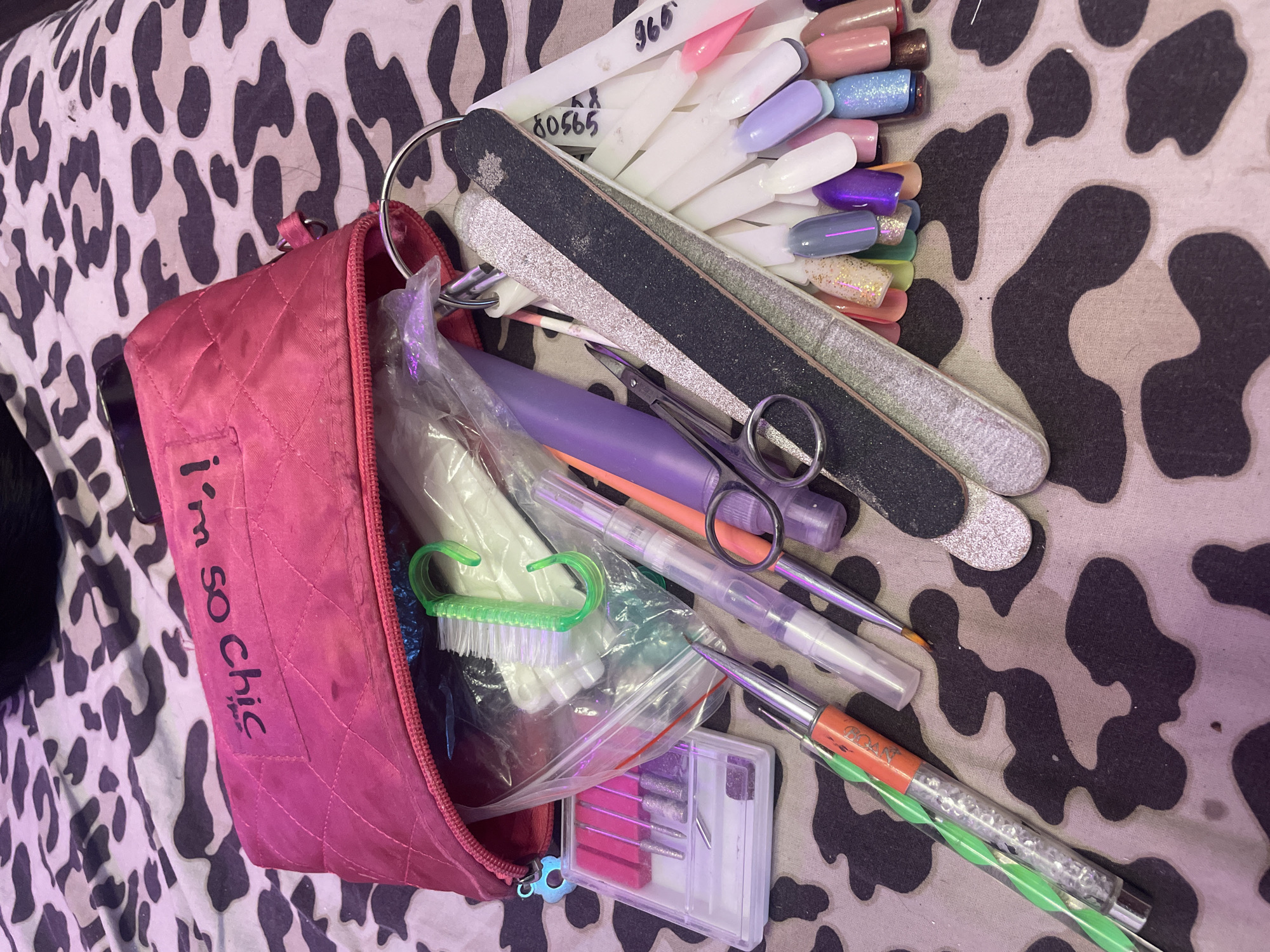 The image displays a collection of nail art supplies scattered on a leopard-print fabric. A pink quilted makeup bag is open, with some contents spilling out. Visible items include nail files of various grits and colors, an assortment of fake nails on display sticks, cuticle scissors, a nail brush, several small tubes and bottles, and a set of drill bits in a clear case. The overall impression is one of organized chaos, suggesting recent use or preparation for nail art activities. The background is out of focus and appears to be a bedspread or similar textile. There is no information about the location in the city of Unknown, Russia.