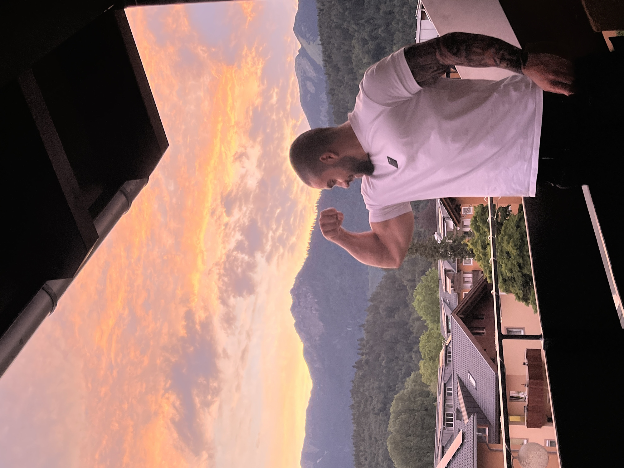 A man is standing on a balcony with a scenic mountain view at sunset. The sky is filled with vibrant orange and pink clouds. The man is flexing his bicep, wearing a white t-shirt and black pants. His arm is tattooed. In the background, there are houses with tiled roofs and lush green trees. The mountains appear in the distance, with some snow on their peaks. The location is Garmisch-Partenkirchen, Germany.