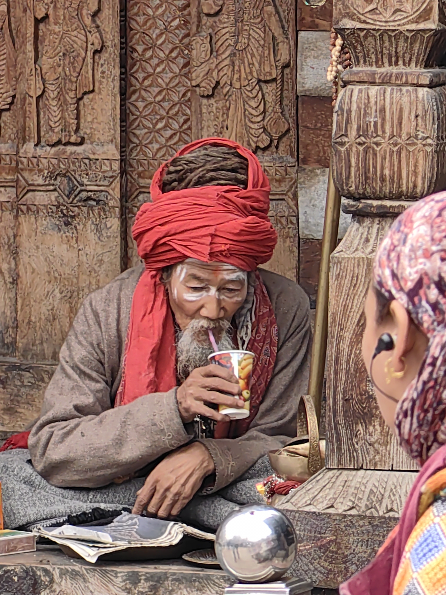 In Vashisht, India, an elderly man with dreadlocks, a red turban, and white face paint is seated and drinking from a cup with a straw. He wears a brown robe and a red scarf. In front of him are some papers and a silver sphere. To his right, another person wearing a colorful headscarf and earbuds is partially visible, facing away from the camera. The background features intricately carved wooden structures, suggesting a spiritual or historical setting.
