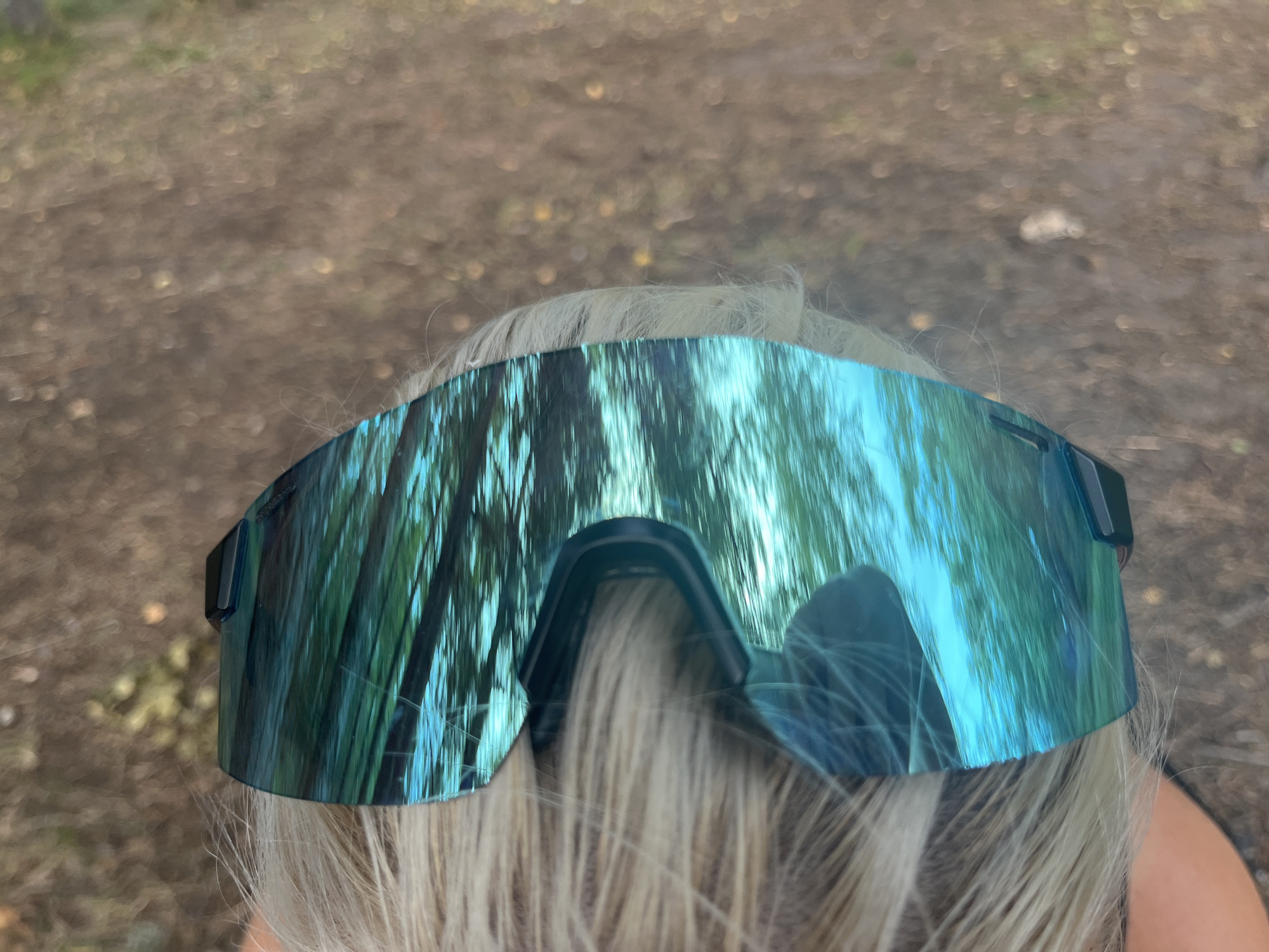 A close-up, overhead shot shows a pair of reflective, teal-tinted sunglasses perched on the blonde hair of a person. The sunglasses are wide and shield-like, with a dark frame. The reflective lenses mirror a scene of tall, thin trees with green foliage, likely in a forest setting. The background is a blur of brown and green, suggesting an outdoor environment. The image appears to be taken at ground level, looking upwards. The location is not specified to be in Pillapalu, Estonia.