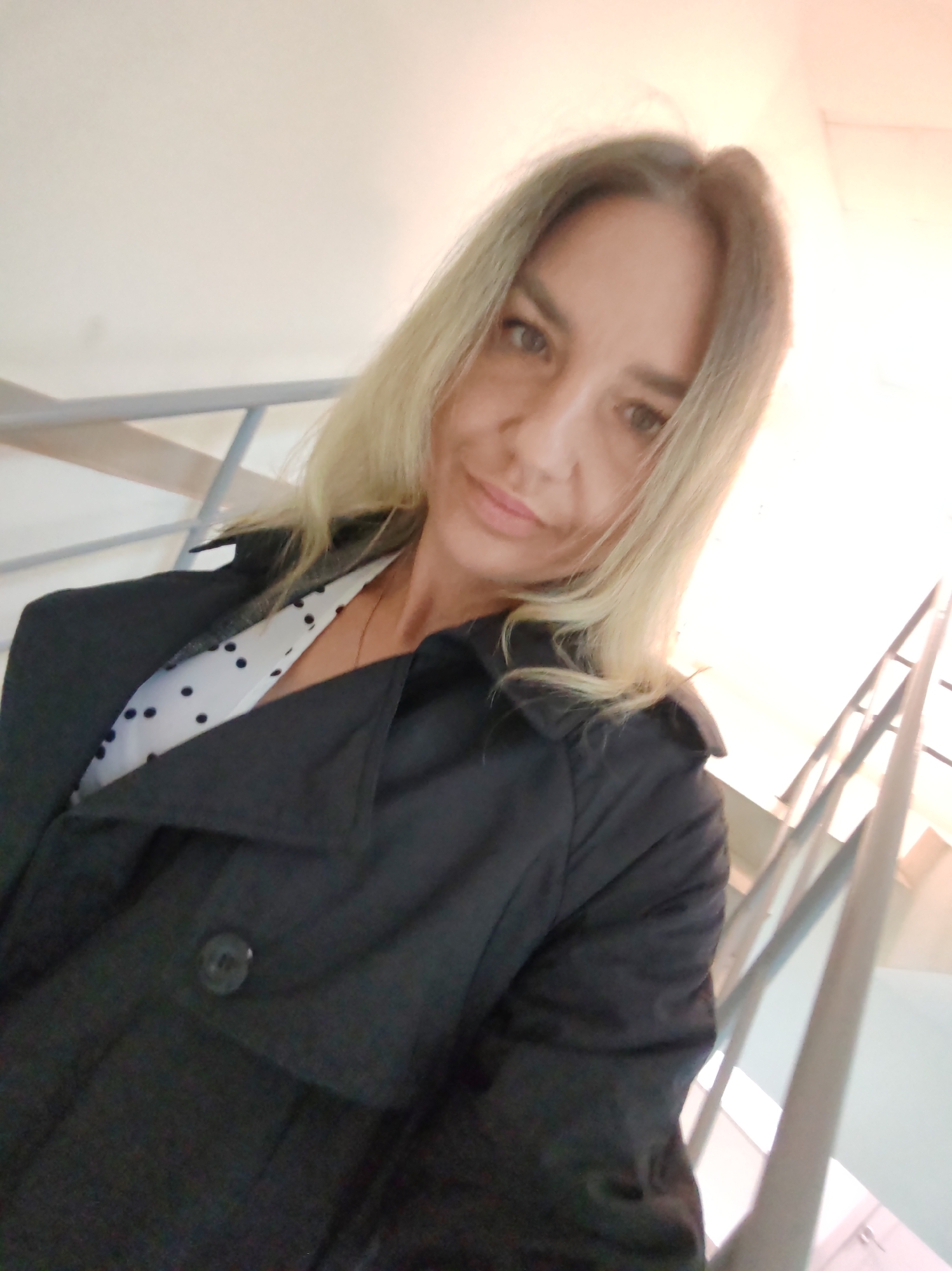 A mid-shot selfie features a woman looking directly at the camera with a gentle smile. She has long, straight light blonde or light brown hair parted in the middle. She appears to be in her 30s or 40s, with a relaxed and pleasant expression.
The woman is wearing a dark, possibly black or navy, coat or jacket, which has a prominent collar and a visible button closure on the front. Underneath, she has on a white blouse or shirt adorned with black polka dots.
The background indicates an indoor setting, likely within a building featuring a staircase or escalator. Sections of light grey or silver metal railings are visible on both sides of the frame, suggesting an upward ascent. The lighting is bright and uniform, typical of an interior space. While the provided context suggests the location is Petropavlovsk-Kamchatsky, Russia, there are no specific visual cues within the image itself to confirm this geographical location. No text is visible in the image.