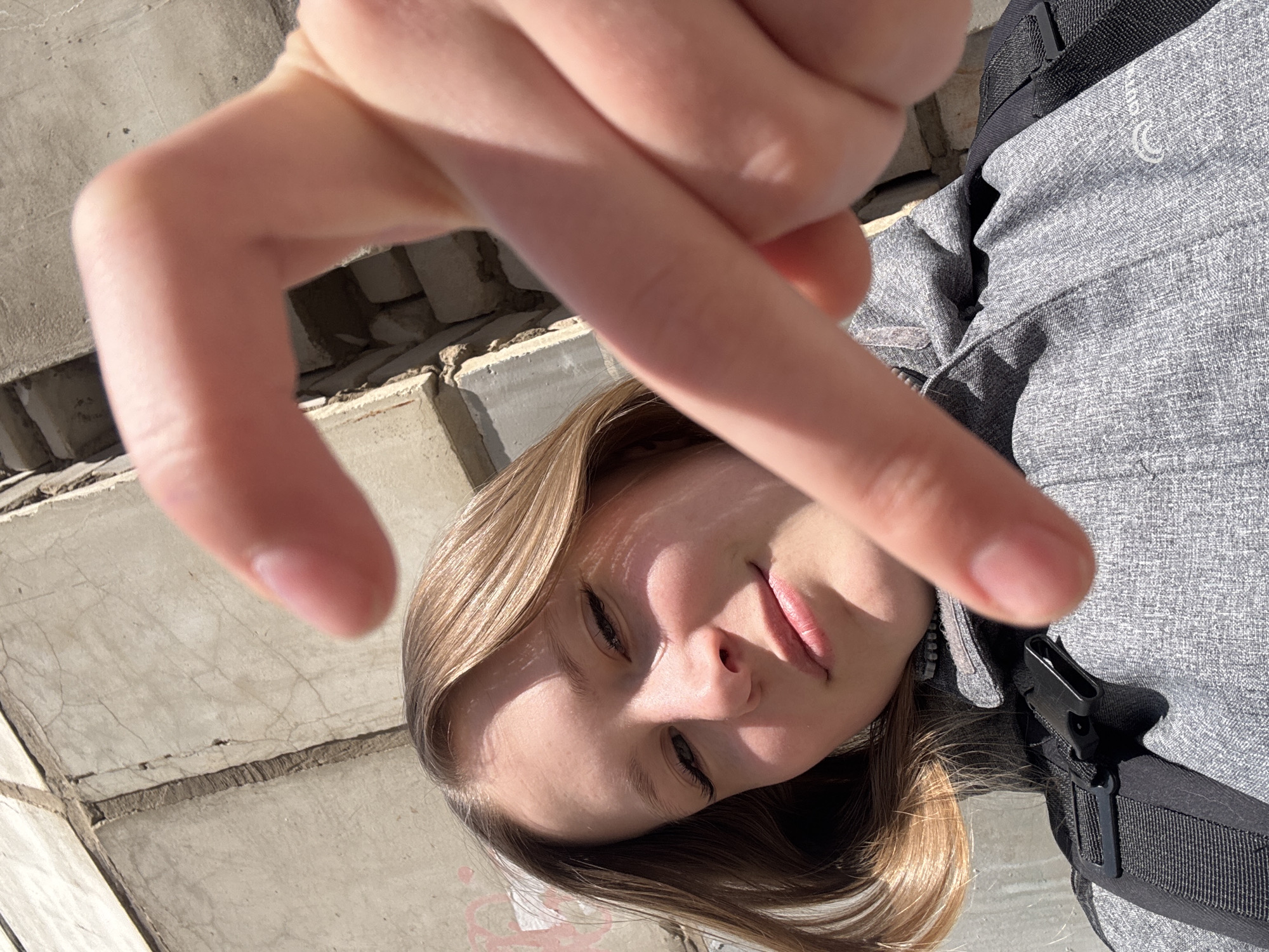 The image, rotated approximately 90 degrees counter-clockwise, presents a close-up, low-angle portrait of a young person in an outdoor setting. A fair-skinned hand, with fingers slightly bent and spread, is prominently featured in the foreground, partially obscuring the subject's face.
The young person has light brown or blonde hair and appears to be looking downwards towards the camera. Their expression is neutral, possibly with a subtle hint of a smile on their lips. Strong, bright sunlight illuminates the scene, casting distinct highlights on the face and hand, and creating shadows that accentuate features.
The subject is wearing a gray, textured jacket, with visible stitching and fabric patterns. A black strap and a black plastic buckle are visible on the right side of the frame, likely belonging to a backpack or a similar item. A small, white, stylized logo, which resembles a "C" or a circular emblem, is visible on the jacket near the shoulder.
The background consists of a weathered concrete or brick structure, showing cracks and signs of age. The bright lighting suggests it is daytime. The overall impression is that of a spontaneous selfie or close-up shot taken from an unusual perspective.