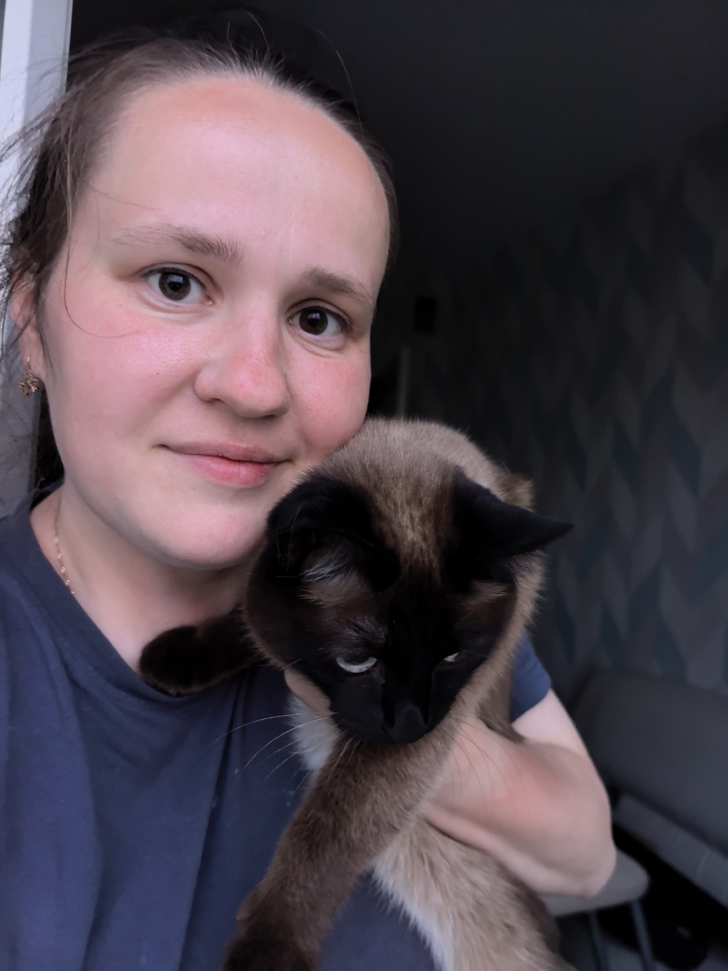 A woman is holding a Siamese cat in an indoor setting. The woman has fair skin, brown eyes, and brown hair pulled back. She is wearing a dark blue shirt and a small gold earring. The cat is predominantly brown with darker brown or black coloring on its face, ears, and paws. The background includes a wall with a geometric pattern in shades of gray and blue. The location is Фролы, Russia.