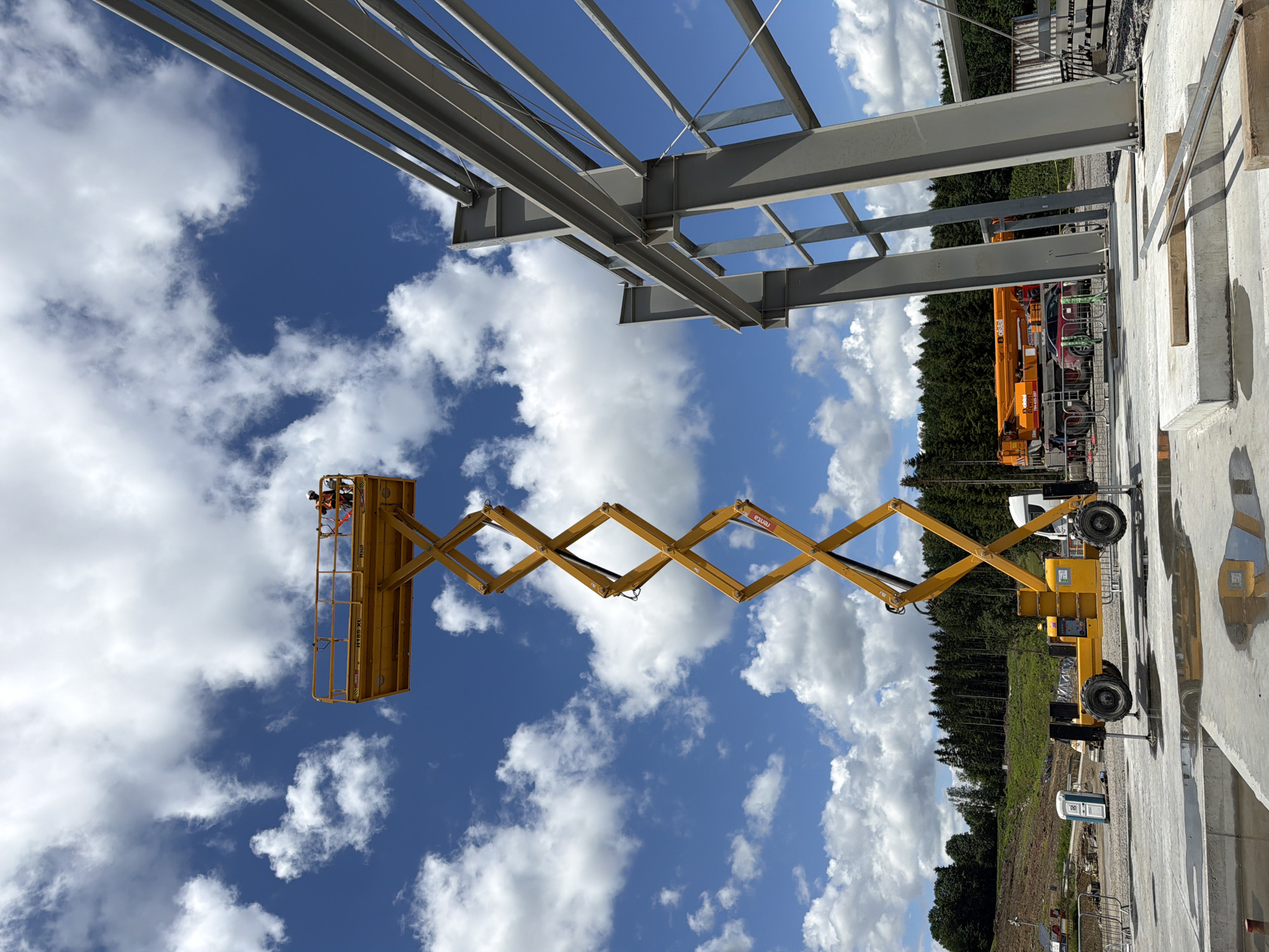 A construction site is depicted under a partly cloudy blue sky. In the foreground, a yellow scissor lift is extended, with one worker wearing a safety harness and orange vest at the top platform. To the right, the steel framework of a building is under construction, with large grey beams. In the middle ground, a portable toilet, a white truck, and an orange crane are visible. The background features a dense forest. The location is identified as Janowice, Poland.