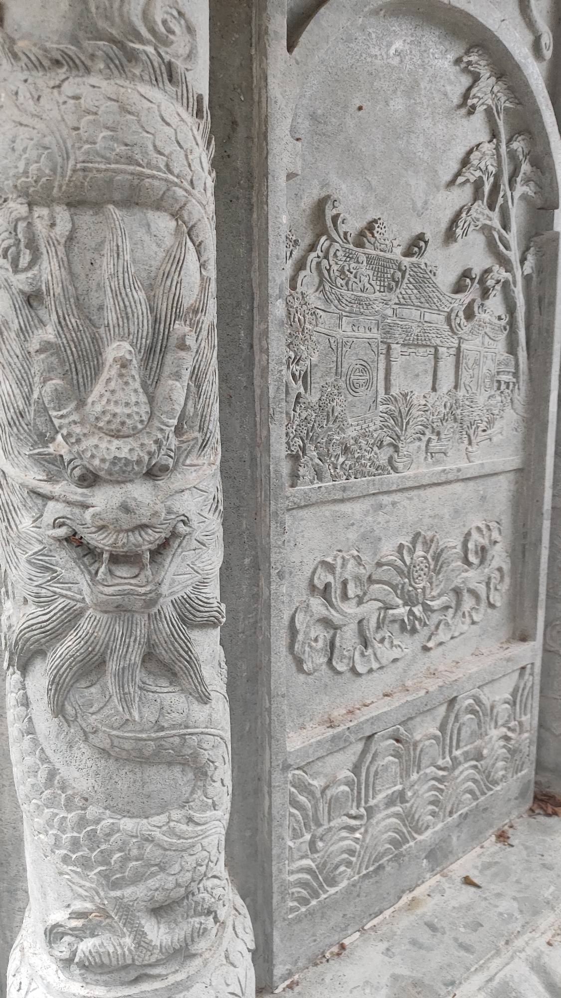 The image shows a detailed stone carving on the exterior of a building, likely a temple or pagoda, in Da Nang, Vietnam. A prominent carved dragon head adorns the left side of the frame, its mouth open in a fierce expression, with elaborate scales and mane. To the right, a carved panel depicts a traditional East Asian building with a tiled roof, surrounded by trees and decorative elements. Below this panel, another carved section features a stylized lotus flower surrounded by swirling motifs. The stonework is weathered, suggesting age and exposure to the elements. The overall style and iconography point to traditional Vietnamese or Chinese architectural ornamentation. The building appears to be constructed from grey stone blocks. The ground in front of the structure is paved with similar stone. Small brown leaves are scattered on the ground and in crevices of the stonework.